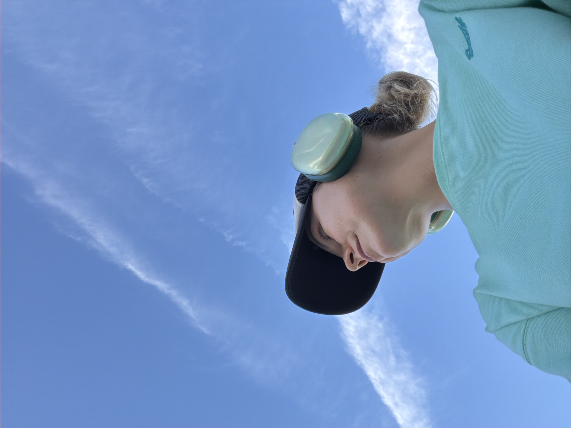 A low-angle shot captures a woman from the chest up, looking upwards into a bright blue sky with wispy white clouds. She is wearing a teal sweatshirt, a black baseball cap, and light green ear protectors. Her hair is pulled back in a bun. The image is taken outdoors, likely in a park or open area, as suggested by the expansive sky and lack of urban elements. The specific location in Nha Trang Ward, Vietnam, cannot be determined from the visual information provided in the image.
