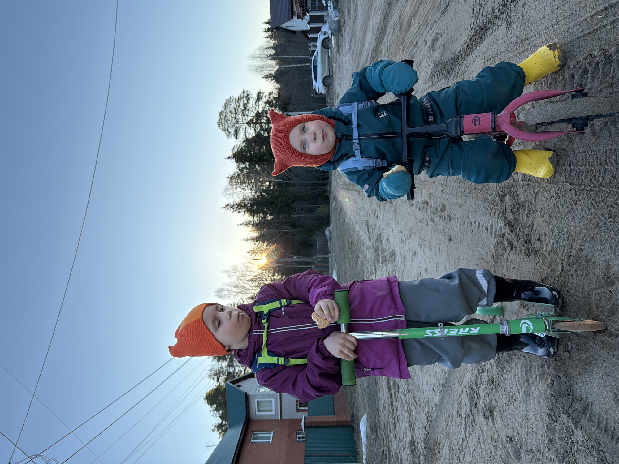 Two children are on a sandy surface outdoors, possibly a construction site or undeveloped area near a residential zone. One child, wearing an orange hat, purple jacket, and gray pants, is standing on a green "KRESS" scooter, holding a cracker. The other child, wearing a red fox-ear hat, a teal jumpsuit, and yellow boots, is riding a pink and black pogo stick. In the background are trees, power lines, a house with a green roof and a brick wall, and a light-colored vehicle. The setting is likely daytime due to the light in the sky. The city is Chaikovsky, Russia.