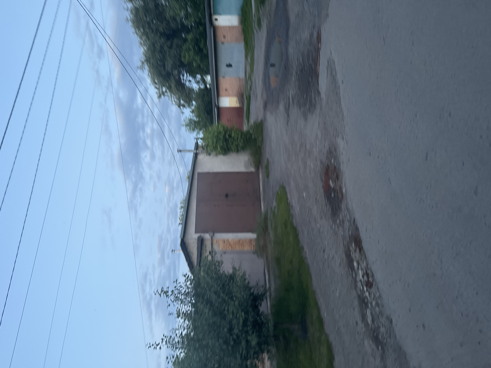 The image depicts a row of garages in the city of Гожули, Ukraine. The garages are constructed of concrete and brick, with corrugated metal doors. Some of the doors are brown, while others are various colors including red, beige, light blue, and white. The area in front of the garages is paved with asphalt, and there are puddles of water on the ground. A tree with green foliage stands to the left of the foreground garage. Power lines are visible in the sky above. The overall impression is one of a somewhat aged and weathered urban environment.