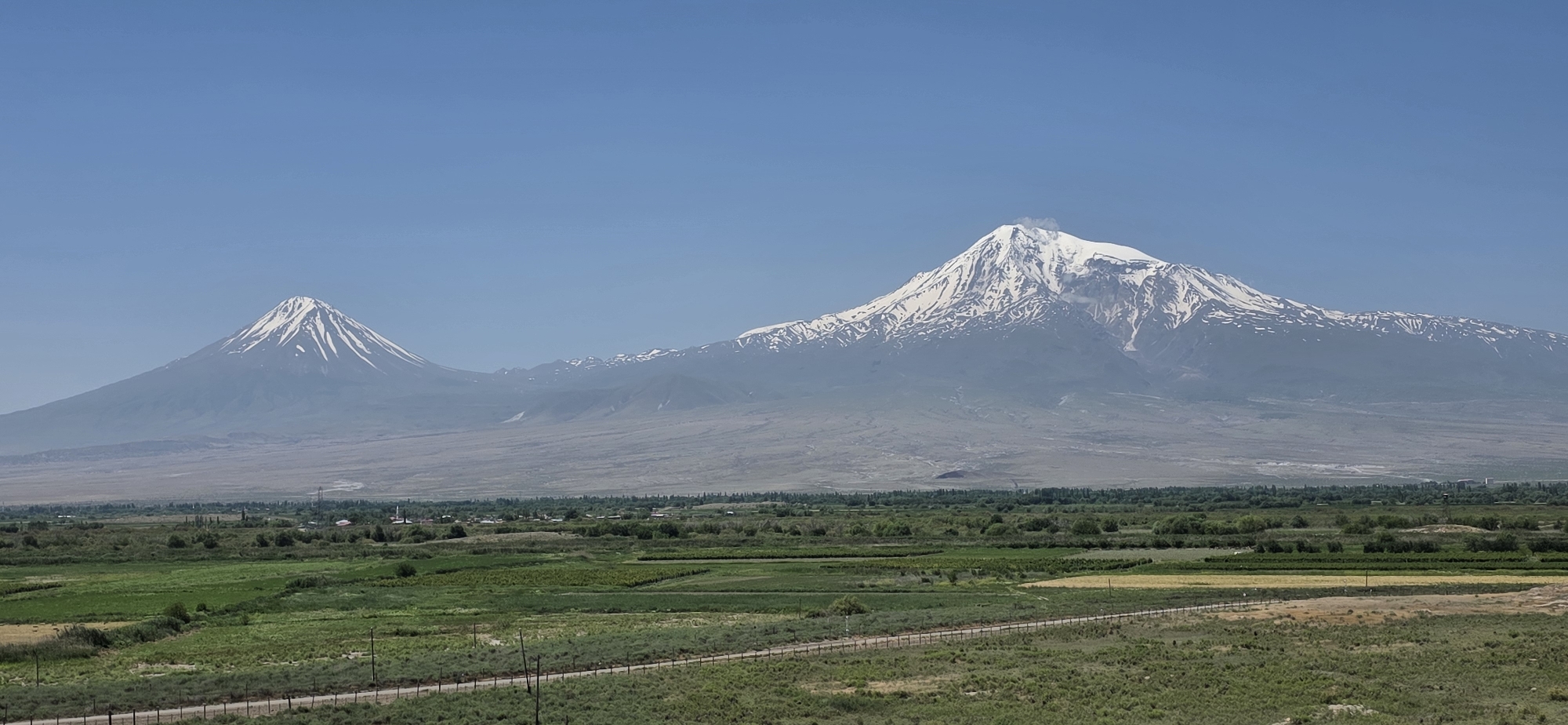 The image depicts a panoramic view of Mount Ararat, a dormant volcano straddling the border between Turkey and Armenia, as seen from the city of Lusarat, Armenia. The foreground is dominated by a vast expanse of green fields and cultivated land, with a dirt road or path running horizontally across the middle ground, bordered by a wire fence. Small clusters of trees and some buildings, indicative of a village or agricultural structures, are scattered throughout the fields. In the background, Mount Ararat rises majestically, its peaks capped with snow. The larger peak, Greater Ararat, is prominent, with a plume of smoke or steam emitting from its summit, suggesting volcanic activity or atmospheric conditions. A smaller, conical peak, Lesser Ararat, is visible to the left of Greater Ararat. The sky is a clear, bright blue. The overall scene conveys a sense of rural tranquility and natural grandeur.