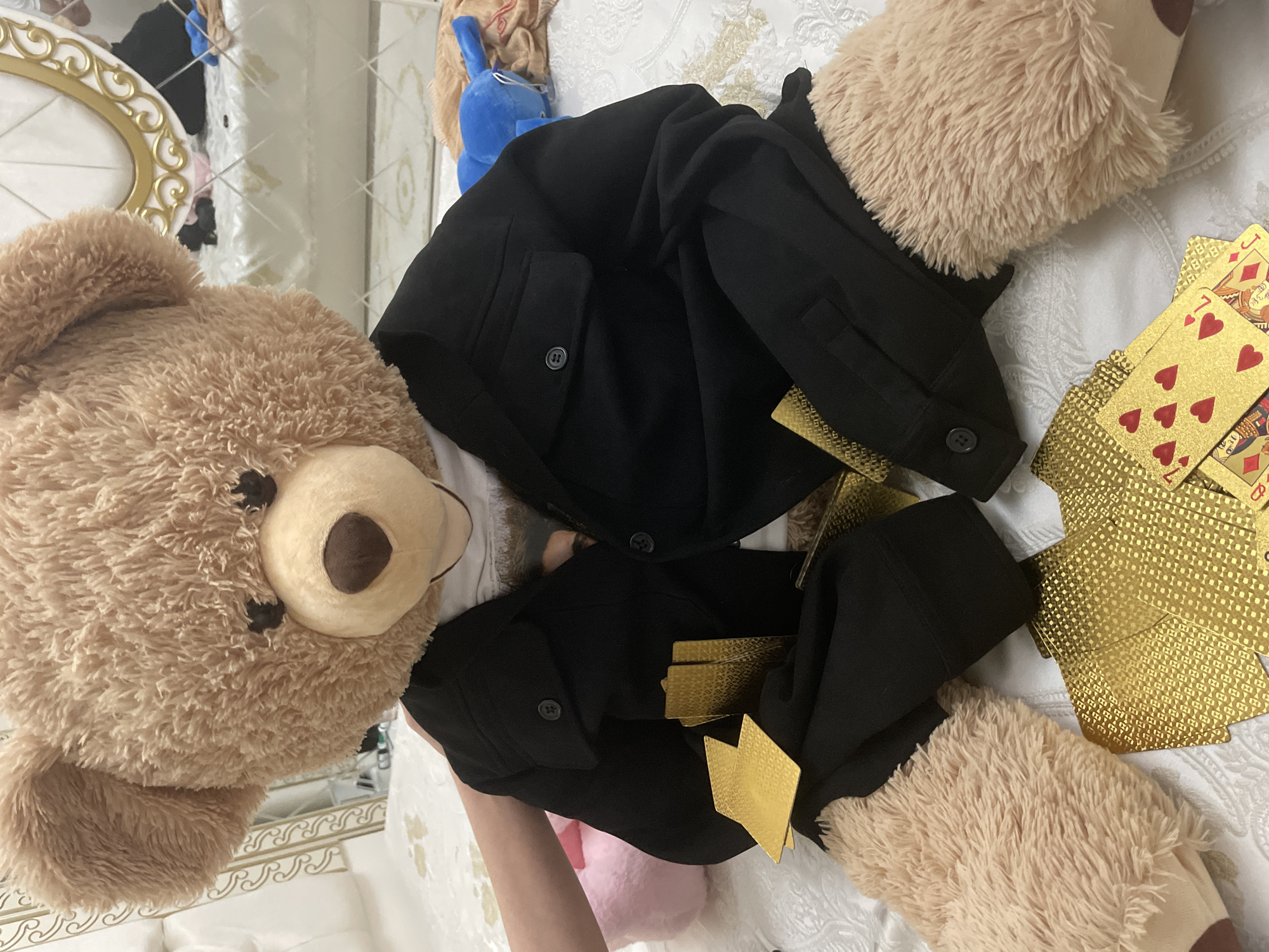 The image presents a playful indoor scene, likely arranged on a bed covered with a white, intricately patterned fabric. The primary subject is a large, light brown teddy bear, positioned prominently on the left side, featuring dark brown eyes and nose. This teddy bear is depicted "wearing" a black jacket or coat, which includes visible buttons and pockets, suggesting a human-like outfit.
Numerous gold-colored playing cards are scattered across the white fabric and around the teddy bear and its jacket. Several cards are face-up, clearly displaying the suit of red hearts, along with traditional card figures such as a King and a Jack, and numeric cards like the 10 of Hearts. The reverse side of the cards features an elaborate, textured gold pattern.
In the upper part of the image, behind the black jacket, a blue plush toy, possibly a rabbit or similar animal, is partially visible. A human hand or arm can be seen at the bottom-left, appearing to adjust or interact with the black garment, indicating human involvement in setting up the scene.
The background elements include reflections from what seems to be an ornate white and gold mirror frame, hinting at a decorative interior space. The lighting is soft and consistent with an indoor environment. The overall impression is one of a whimsical, staged moment, portraying the teddy bear as if it's participating in a card game or a similar imaginative scenario. No specific visual cues are present in the image to confirm the location context of Ihtiman, Bulgaria.