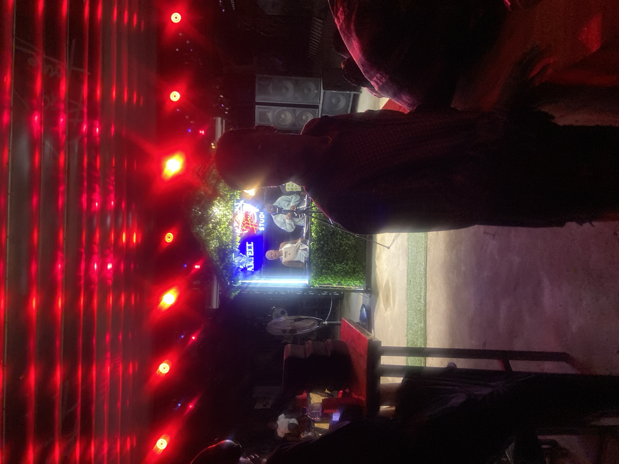 In Howayi, Nigeria, an indoor event is underway, illuminated by an abundance of red lights mounted on the ceiling and on a structure that resembles a booth. The booth is decorated with artificial greenery and features a neon sign that reads "ARK TELL STUDIO". Within the booth, two individuals are visible: one is standing and appears to be interacting with equipment, while another is behind a counter, smiling. In the foreground, a person is seated, silhouetted against the ambient lighting. A red table with stacked cushions is positioned to the left, and a standing fan is also present. In the background, large speaker cabinets are stacked, and other individuals are partially visible. The overall atmosphere suggests a lively social gathering or a performance space.