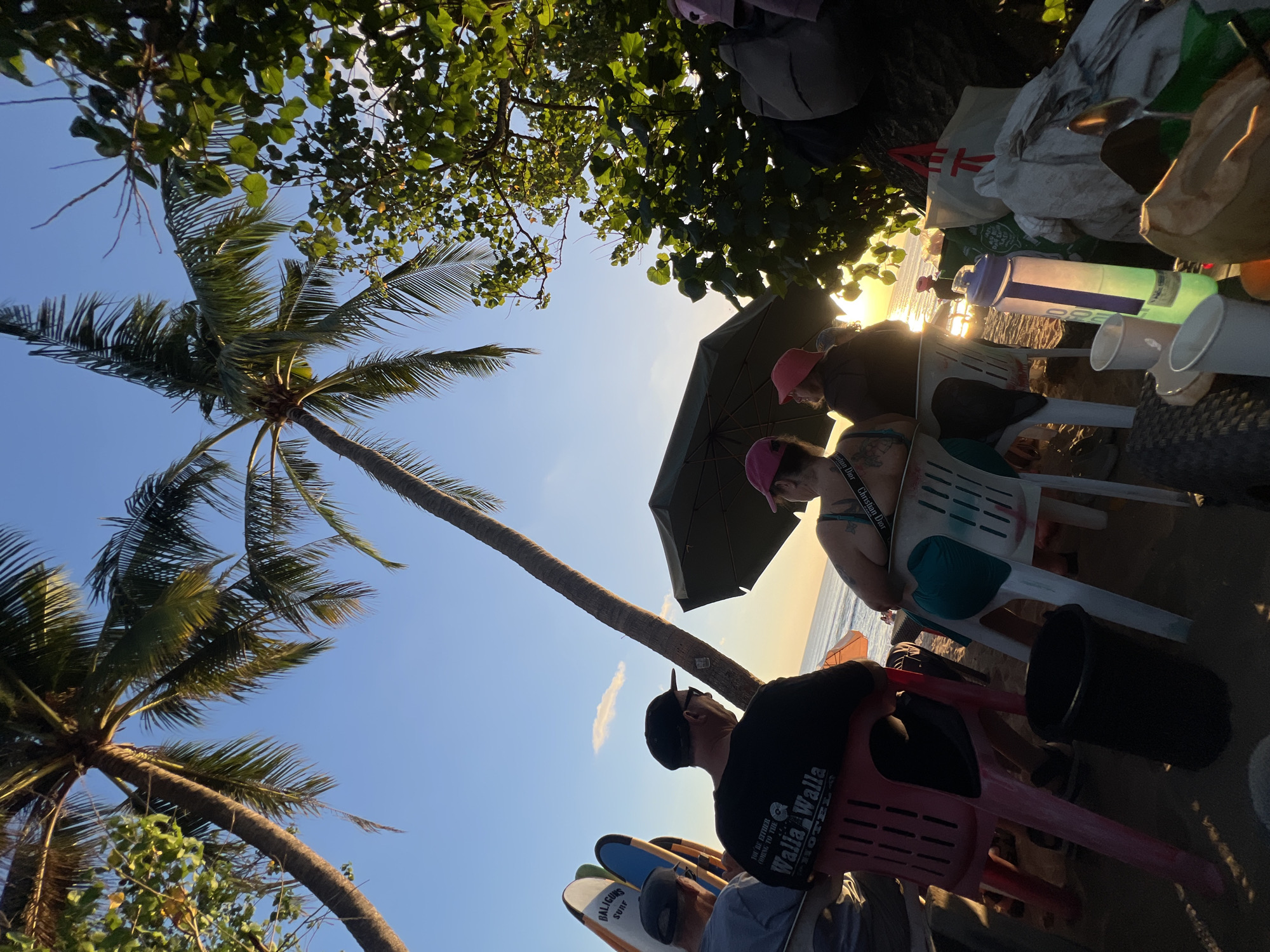The image depicts a beach scene in Kuta, Indonesia, during sunset. In the foreground, several people are seated on plastic chairs on the sand, facing the ocean. A large green umbrella provides shade. Palm trees are visible, silhouetted against the bright sky.
Among the people, one individual is wearing a black t-shirt with white lettering that reads "Walla Walla HOTEL". Another person has a bright blue design on their clothing. To the left, several surfboards are propped up.
In the middle ground, the ocean and the setting sun are visible, casting a warm glow on the water and sky. A few other people can be seen in the distance on the beach.
The overall atmosphere is relaxed and casual, suggesting a group enjoying a sunset by the sea. The presence of surfboards indicates the location is likely a popular spot for surfing. The lighting suggests it is late afternoon or early evening.
