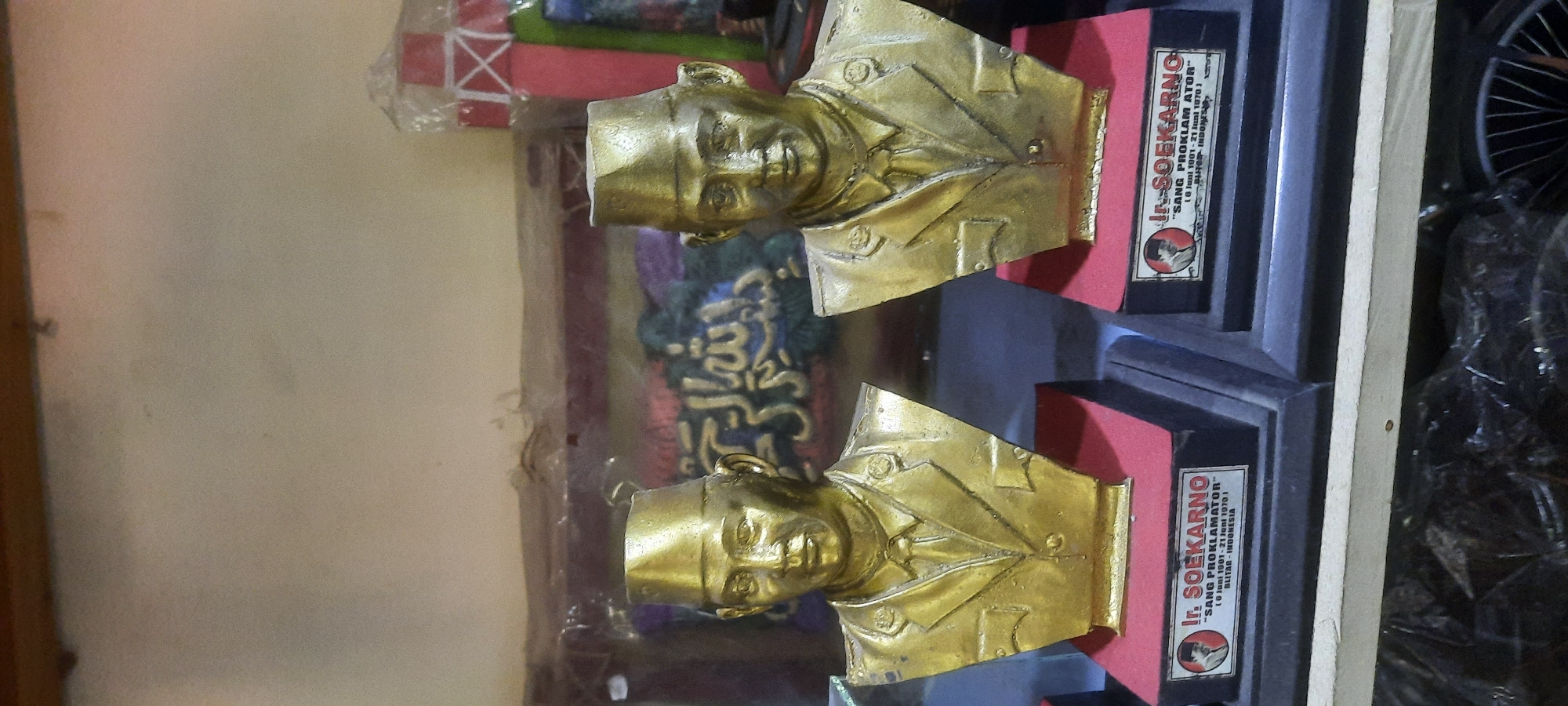 The image displays two golden busts of Ir. Soekarno, positioned side-by-side on dark bases with red surfaces. Each bust is mounted on a rectangular plaque with text and a small portrait of Soekarno. The plaque on the left reads "Ir. SOEKARNO" with "SANG PROKLAMATOR" and dates "[6 Juni 1901 - 21 Juni 1970]" and "BLITAR - INDONESIA" below. The plaque on the right is partially obscured but appears to have identical text. In the background, there's a blurred tapestry with Arabic calligraphy, suggesting a cultural or religious context. The overall scene is indoors, with the busts likely displayed on a shelf or table.
