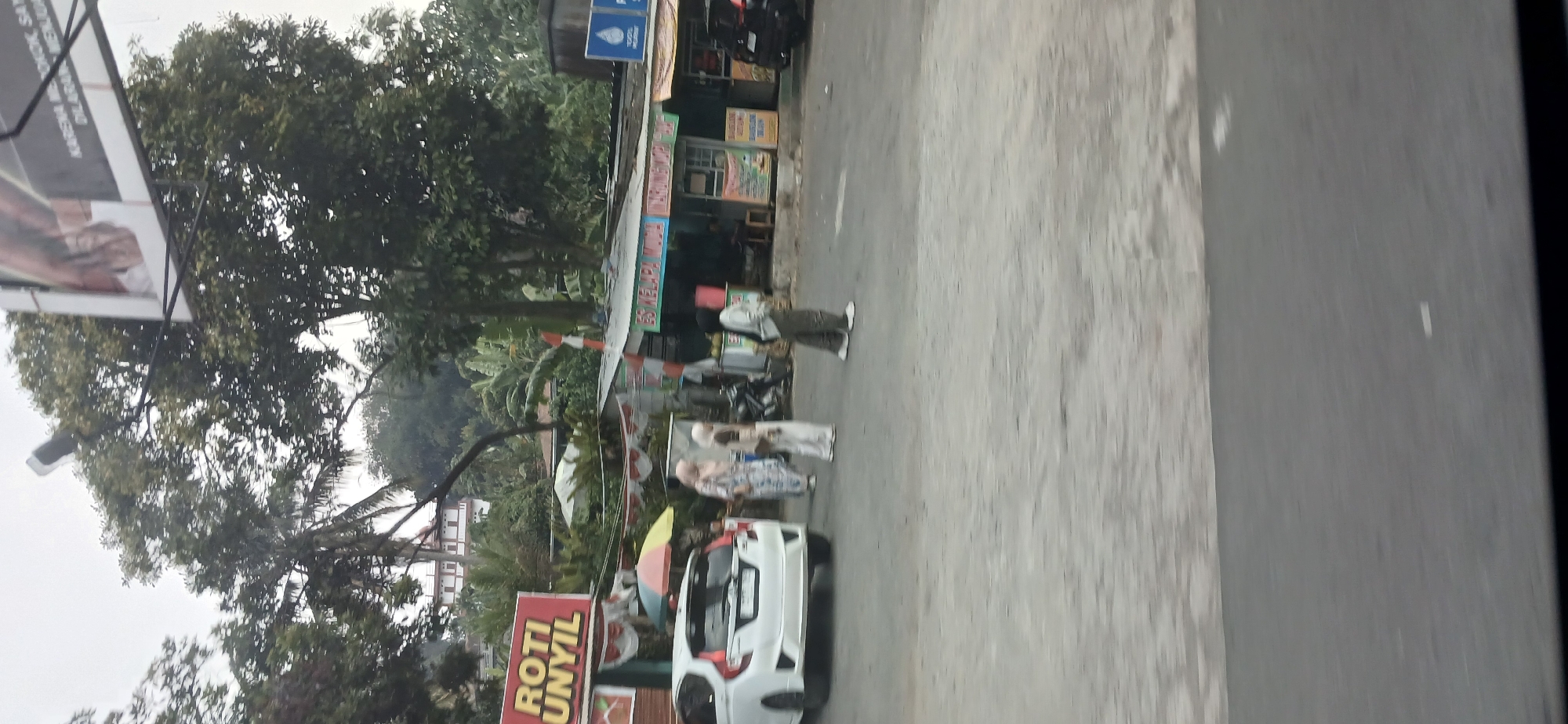 The image depicts a street scene in Ciawi, Indonesia, with a focus on a roadside food stall. On the left, a white car is parked. In the foreground, three individuals are visible. One person is wearing a white hijab and a patterned dress, another wears a beige hijab and a white outfit, and a third person in the background is wearing a light-colored hijab and pants. Behind the individuals are several food stalls with various signs. One sign prominently reads "ROTI UNYIL". The stalls are adorned with Indonesian flags and decorations. Trees and lush vegetation are abundant in the background, with a multi-story building visible on a hill. The sky is overcast, suggesting a cloudy day.