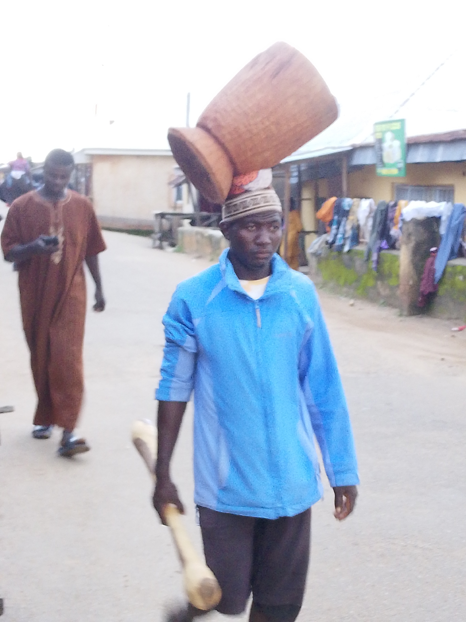 In a daytime scene on a dusty street in Jos, Nigeria, a young Black man, appearing to be in his late teens or early twenties, walks forward, his face turned slightly to the left. He wears a blue zip-up jacket over a yellow t-shirt and dark trousers. A traditional cap with geometric patterns sits on his head, and a large, hollow, wooden object, possibly a drum or a large container, is balanced precariously on top of the cap and his head. In his right hand, he holds a light-colored wooden stick or drum beater.
In the background, another Black man, older, walks away from the camera, wearing a long, brown, traditional robe. He holds a mobile phone in his left hand. Further in the background, buildings with corrugated metal roofs line the street, and clothes hang on a line or fence, suggesting a residential or market area. The ground is unpaved and dusty. The sky is overcast and bright. The overall impression is of a busy, possibly everyday, moment in an urban Nigerian setting.