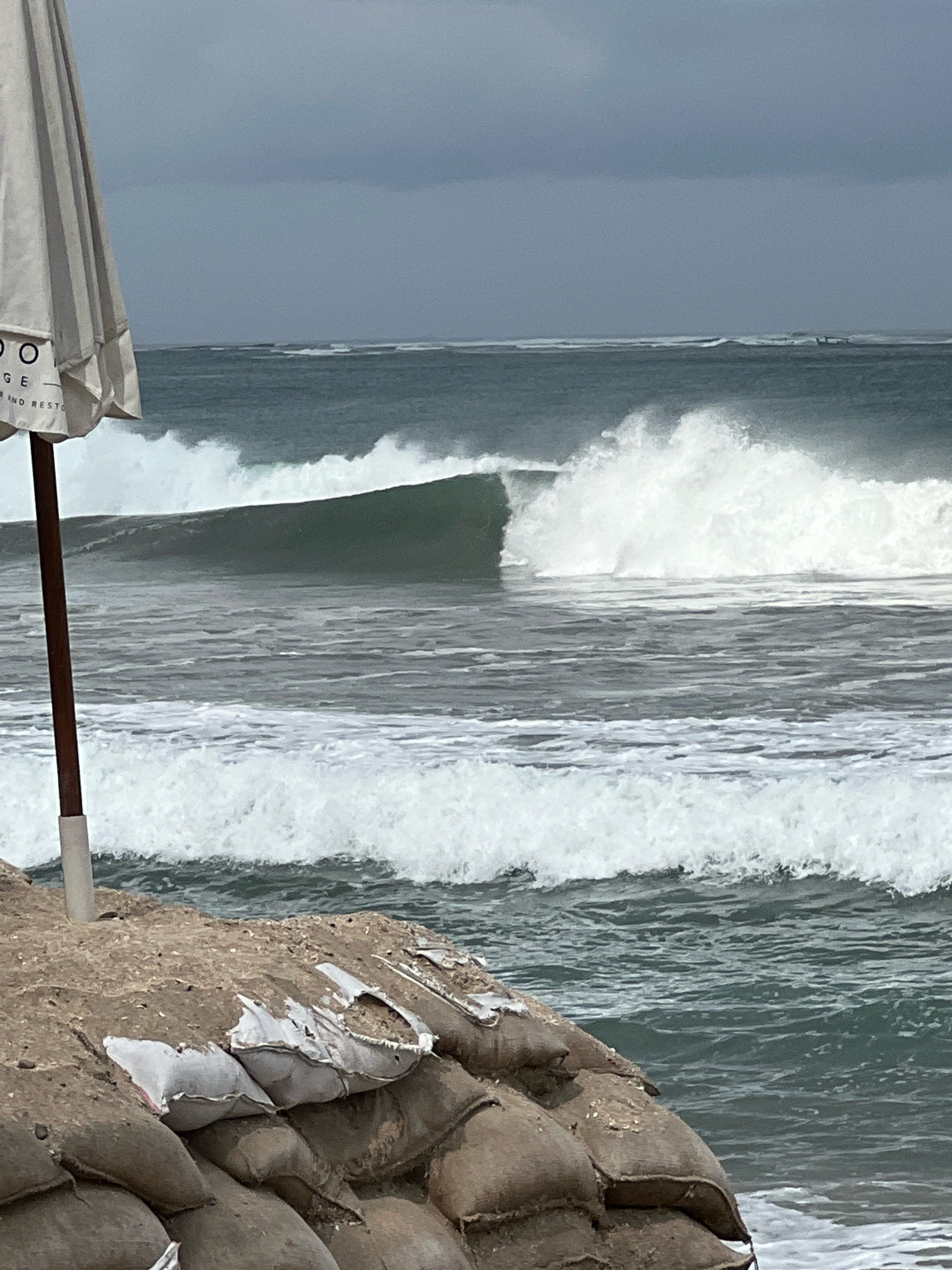 A stormy, overcast sky looms over Kuta Beach, Indonesia, where a large umbrella stands prominently in the foreground, partially obscuring a view of the powerful ocean. The umbrella is white with black lettering, and its fabric is taut, indicating a windy day. In front of the umbrella, a wooden pole supports it, and a stack of sandbags, some torn and spilling their contents, lines the beach. These sandbags are a dark, sandy color and are piled high, suggesting a measure against erosion or flooding.
The ocean is the main subject of the midground, with large, dark green waves breaking and crashing. One particularly large wave is captured in mid-curl, its crest a frothy white. Smaller waves and whitewater are visible further out to sea and closer to the shore. In the far distance, a faint line of waves suggests a consistent swell, and a few tiny figures, possibly surfers or small boats, can be discerned against the horizon. The water is a deep, choppy gray-green, reflecting the somber sky. The overall impression is one of a dramatic and wild coastal scene, typical of stormy weather on a popular Indonesian beach.