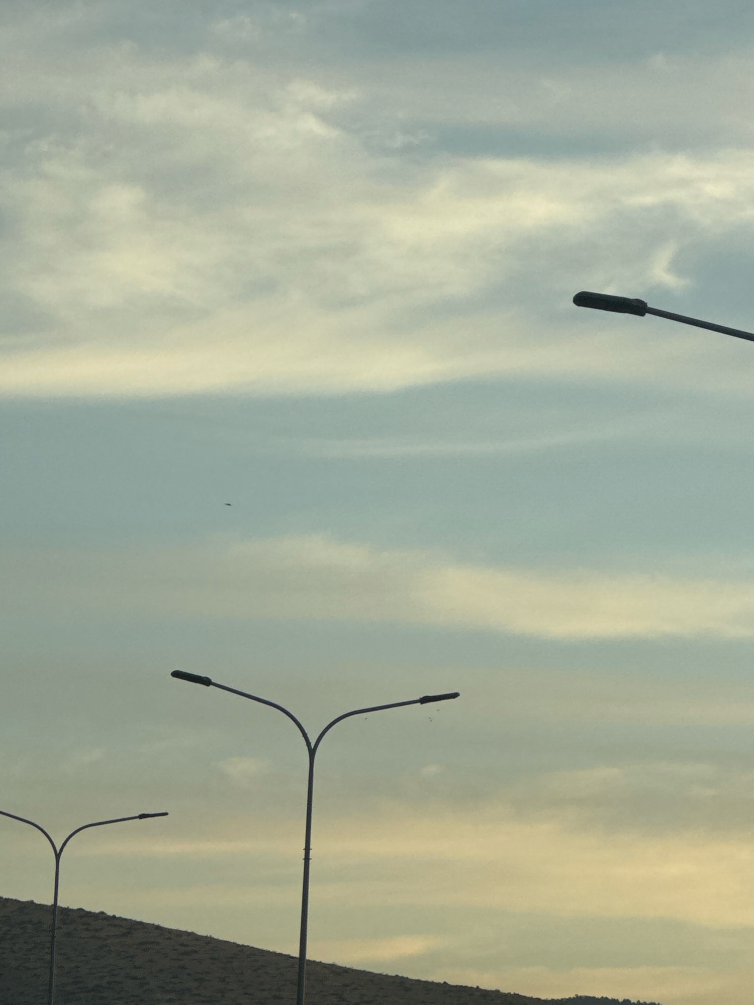 The image captures a cloudy sky with wispy, horizontal cloud formations in shades of light blue and pale yellow. Three lampposts are visible in silhouette against the sky. The lampposts are modern, with curved arms supporting dark, elongated light fixtures. Two lampposts are in the foreground, one on the left and a taller one to its right, both positioned over a dimly lit, undulating landscape that appears to be a hillside or embankment covered in vegetation. A third lamppost is positioned higher and further to the right, with only its light fixture and arm visible against the sky. A small, dark object, possibly a bird or an insect, is visible in the upper middle section of the sky. The scene suggests an outdoor setting, likely along a road or in a public area, during either dawn or dusk due to the lighting conditions. The location is identified as Germasogeia Municipality, Cyprus.