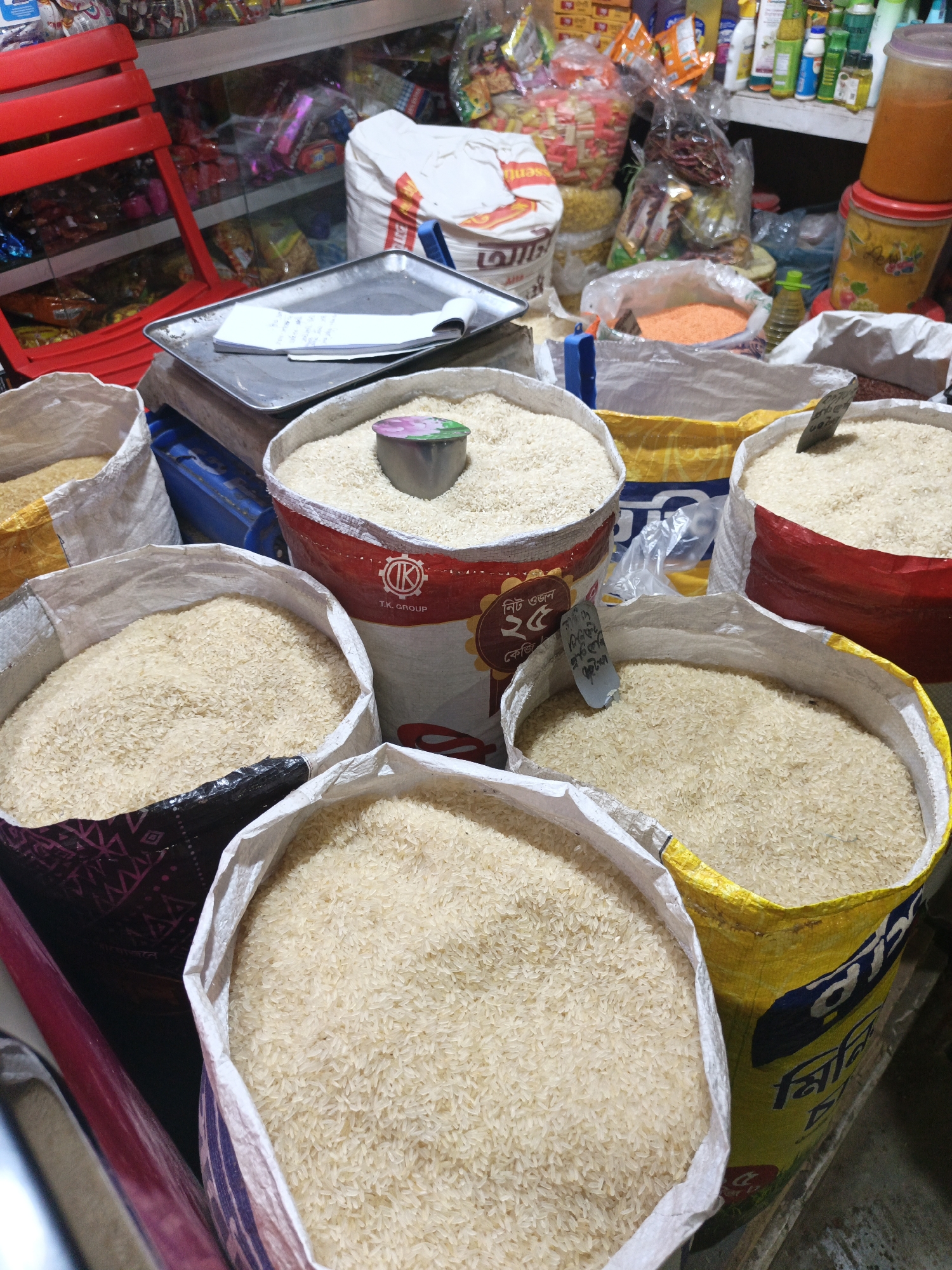 The image depicts a market setting, potentially in a shop or a section of a larger marketplace. There are multiple sacks or bags filled with grains, most likely rice, arranged closely together. The bags have different designs and colors, including red, yellow, and black patterns, suggesting variety in the type or brand of rice being sold. A metal scoop rests in one of the bags, indicating that the grains are available for purchase in specific quantities.
In the background, a variety of other products are visible on shelves, including food items, household goods, and possibly personal care products. A weighing scale with a notebook on top is also present, suggesting the method of measuring and recording sales.
The setting appears to be a busy commercial area, likely in a city. Due to the language on the bags and signage, the location is possibly in Bangladesh.