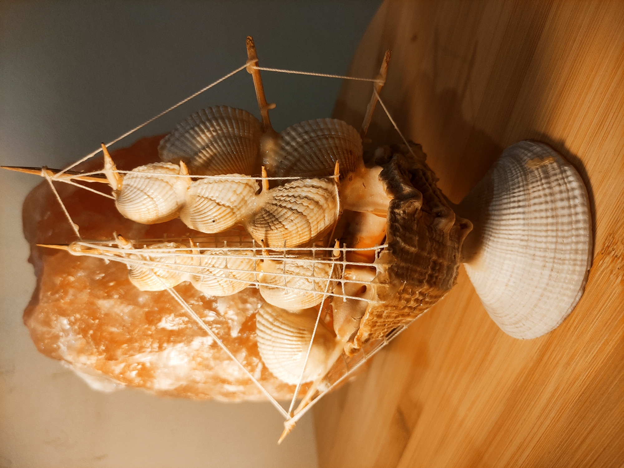 A decorative model ship constructed of seashells and string sits atop a salt lamp, with both objects resting on a wooden surface. The ship features several ribbed, white shells used as sails, attached to a frame of thin wooden sticks and secured with white string. A larger, textured brown shell forms the stern, while another white shell is positioned at the rear. The salt lamp, a solid mass of pinkish-orange crystal, supports the ship from below. The backdrop is a gradient from a pale beige wall to a medium brown wooden surface.