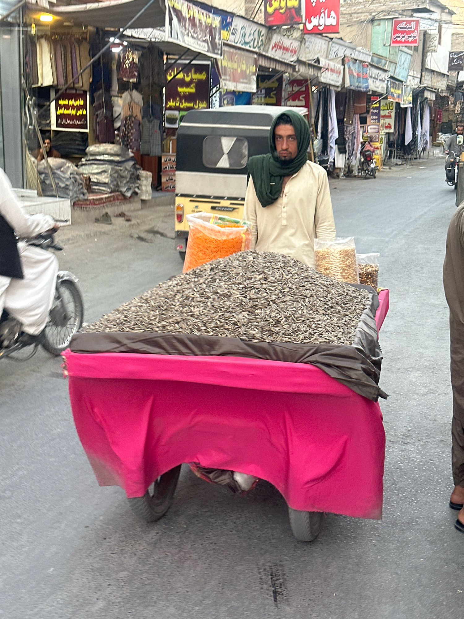 The image depicts a street scene in Quetta City Tehsil, Pakistan, likely a marketplace given the stalls and vendors. In the foreground, a man wearing a green headscarf and a light-colored tunic stands behind a cart laden with a large pile of sunflower seeds. The cart is covered with a vibrant pink cloth. To the left, a person on a motorcycle is visible, wearing white attire. In the background, various shops line the street, displaying clothing and other goods. Signs with text in Urdu are prominent on the shop fronts. A three-wheeled vehicle, possibly a rickshaw, is also present. The overall environment suggests a bustling urban setting with commercial activity.