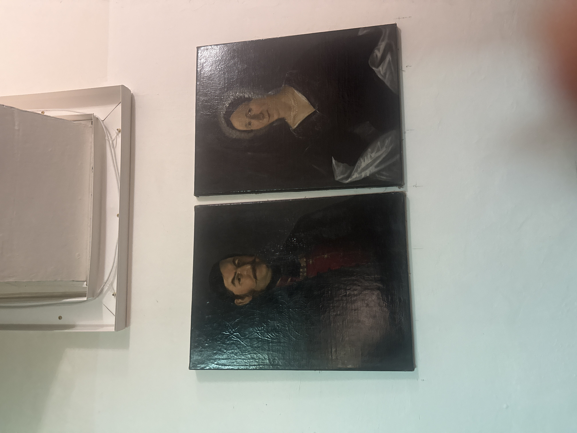 The image shows two paintings hung on a white wall, alongside a square, white box-like structure with exposed screws and internal wires. The top painting depicts a woman, viewed from the shoulders up, wearing a dark gown with a silver collar, set against a dark background. The bottom painting shows a man, also from the shoulders up, in a dark uniform with red detailing, similarly set against a dark background. The wall behind them is plain white. The paintings appear to be affixed to the wall using nails or tacks, visible as small dark spots above each painting. The location is likely Karpaty, Ukraine.