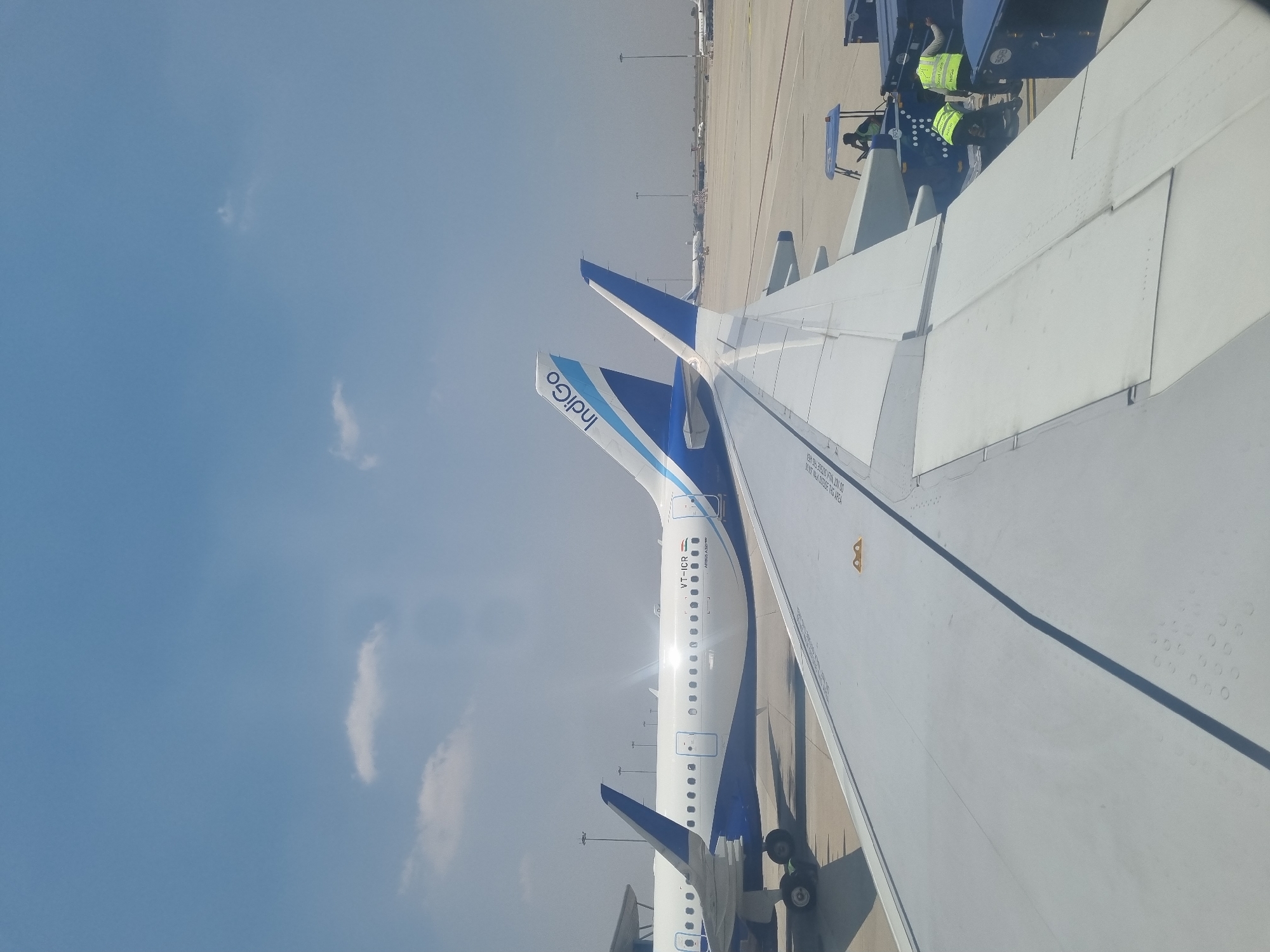 The image captures a view from inside an airplane, looking out over the wing. Another Indigo airplane is visible on the tarmac, with its tail and Indigo logo clearly displayed. Airport ground personnel in bright yellow safety vests are positioned around a luggage conveyor. The sky is blue with some clouds. The tail of the plane says it is VT-ICR. The tarmac is made of cement.
