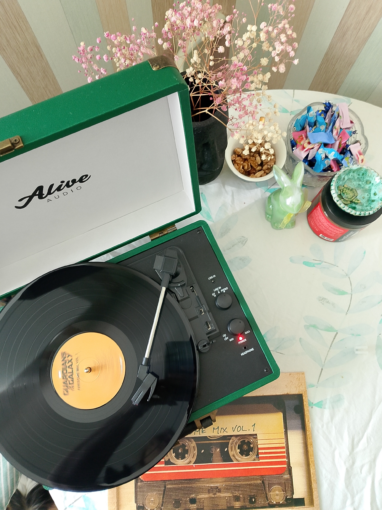 The image presents an overhead view of an indoor tabletop arrangement. A green "Alive AUDIO" portable turntable, housed in a suitcase-style case, is open and positioned in the left-central area. A black vinyl record, labeled "GUARDIANS OF THE GALAXY AWESOME MIX VOL. 1" on an orange center, rests on the platter, with the tonearm extended over it. The turntable's control panel displays inputs for "LINE IN" and "PHONO", a volume control from "MIN" to "MAX", an "ON/OFF" switch with a red "POWER" indicator light, and a "HEADPHONE" jack.
The table surface is covered by a light-colored tablecloth adorned with light green leaf patterns. Various items are placed on the tablecloth to the right of the turntable. These include a dark vase holding dried pink and white baby's breath flowers, a small white bowl containing shelled walnuts, and a clear glass bowl filled with individually wrapped candies in blue, pink, and light green wrappers. A glossy light green rabbit figurine, featuring a yellow ribbon, is situated near these bowls. Further right, a red cylindrical container with a black lid has a small, textured teal-colored dish placed on top of its lid, which contains a miniature golden turtle figurine. Below the turntable, a rectangular object depicts a vintage audio cassette labeled "AWESOME MIX VOL. 1". The background shows a wall with vertical stripes of alternating light green, blue, and beige colors. No individuals are present in the scene. The specific location within Izhevsk, Russia, cannot be determined from the visual information provided.