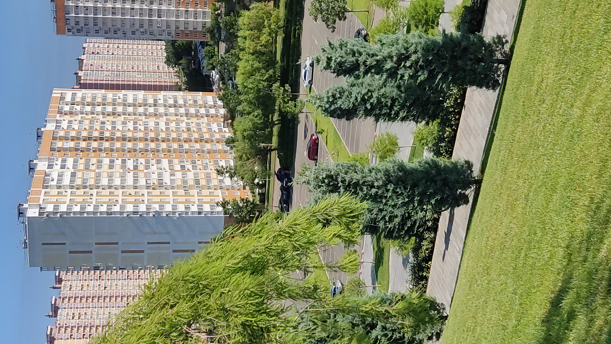 This is a high-angle, wide shot of a modern urban environment on a sunny day. The scene is dominated by tall, multi-story residential buildings with numerous windows and balconies, suggesting a densely populated area. These buildings are lined with trees and manicured green spaces, including a grassy lawn and a paved walkway. A parking lot is visible, with several cars parked and one in motion. The architecture appears to be contemporary, with light-colored facades and repeating patterns of windows. The time of day is likely daytime, given the bright sunlight and the clear blue sky. The location appears to be Krasnodar, Russia, based on the provided context. No people are clearly discernible in the image, but their presence is implied by the cars and residential buildings. The overall impression is one of a well-developed urban neighborhood with ample green spaces.