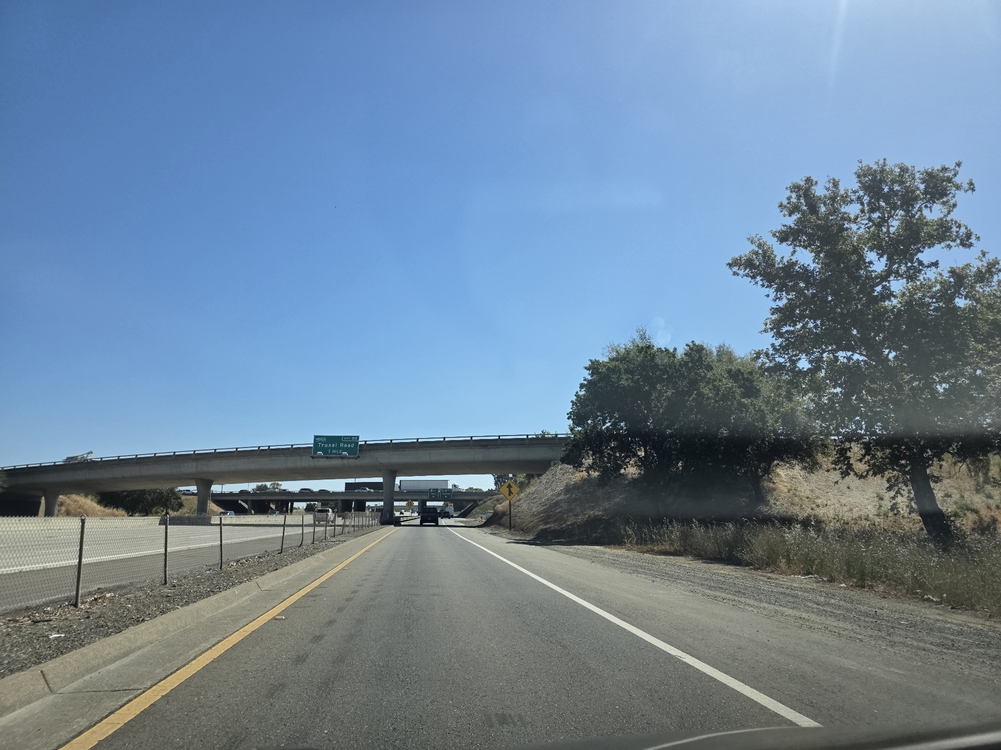 This media file captures a view from the driver's perspective on a highway in Sacramento, California. The scene is set on a clear, sunny day with a bright blue sky, suggesting it is daytime. The highway has multiple lanes, with a concrete barrier and chain-link fence lining the left side.
A prominent overpass stretches across the highway, with traffic visible on it. A green road sign on the overpass indicates "SOUTH I-80 TRUXEL ROAD 1 MILE". To the right of the highway, a dry, grassy embankment is topped with trees, casting shadows on the ground, hinting at a warm climate. A single tree with a broad canopy stands near the embankment. The overall impression is one of travel on a typical sunny day in a semi-arid or Mediterranean climate. No people are clearly visible. The activity is vehicular travel on a highway.