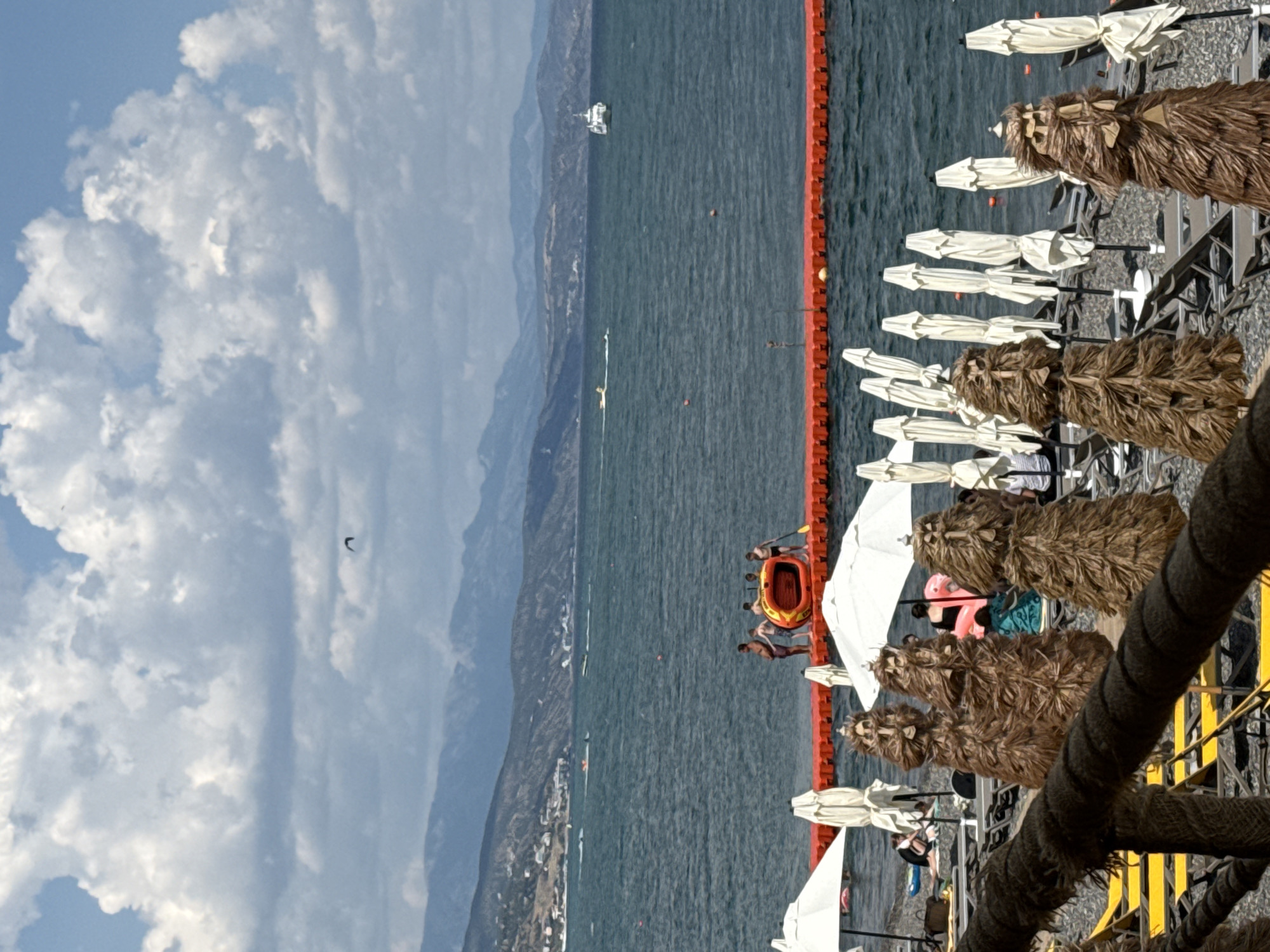 On a sunny day, a wide view of the Black Sea from the coast of Alushta, Ukraine, unfolds. The sea, a deep blue, is dotted with a few small boats and a distant white boat. The shore is lined with a series of white beach umbrellas, some open and some closed, and lounge chairs. In the foreground, several unusual, tall, brown, straw-like figures stand on the beach, resembling scarecrows or artistic installations. A bright orange inflatable raft with people in it is visible in the water, positioned on a floating orange barrier. In the background, a range of hazy blue mountains forms the horizon, with a few scattered buildings visible on the lower slopes. The sky above is a mix of bright blue with large, fluffy white clouds.