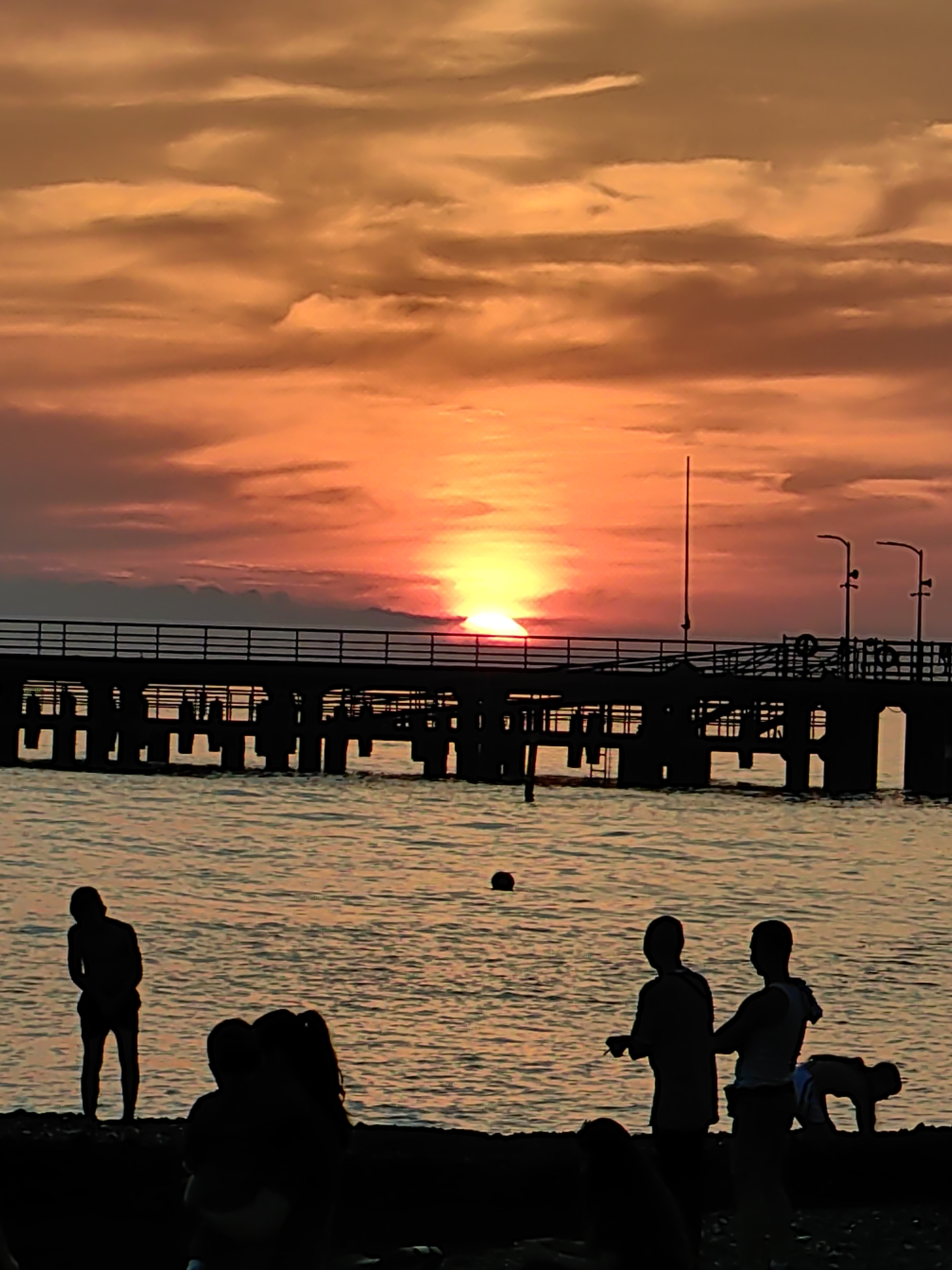 This image depicts a sunset scene in Sochi, Russia, with the sun low on the horizon, casting a vibrant orange, red, and yellow glow across a cloudy sky. The sun's disk is partially obscured by clouds and appears to be just above a long, dark pier that extends across the middle ground.
The pier, silhouetted against the bright sky, features a railing and numerous support pillars submerged in the water. Several streetlights are visible on the pier's surface, along with a vertical pole structure and possibly a lifebuoy. The water in the foreground reflects the warm tones of the sky, showing gentle ripples. A dark, spherical buoy floats in the water to the left of the pier.
On the dark shoreline in the immediate foreground, several human figures are silhouetted. From left to right:
* A solitary person stands facing right, with their back to the viewer.
* Two individuals are seated or crouched together, largely obscured.
* Two more people stand to the right, one appearing to be holding something or gesturing, and the other facing them.
* Further to the right, another person is in a bent-over or crouching position, possibly engaged in an activity on the sand or near the water's edge.
The overall scene captures the tranquil moment of a sunset over the Black Sea, with leisure activities on the shore and pier.