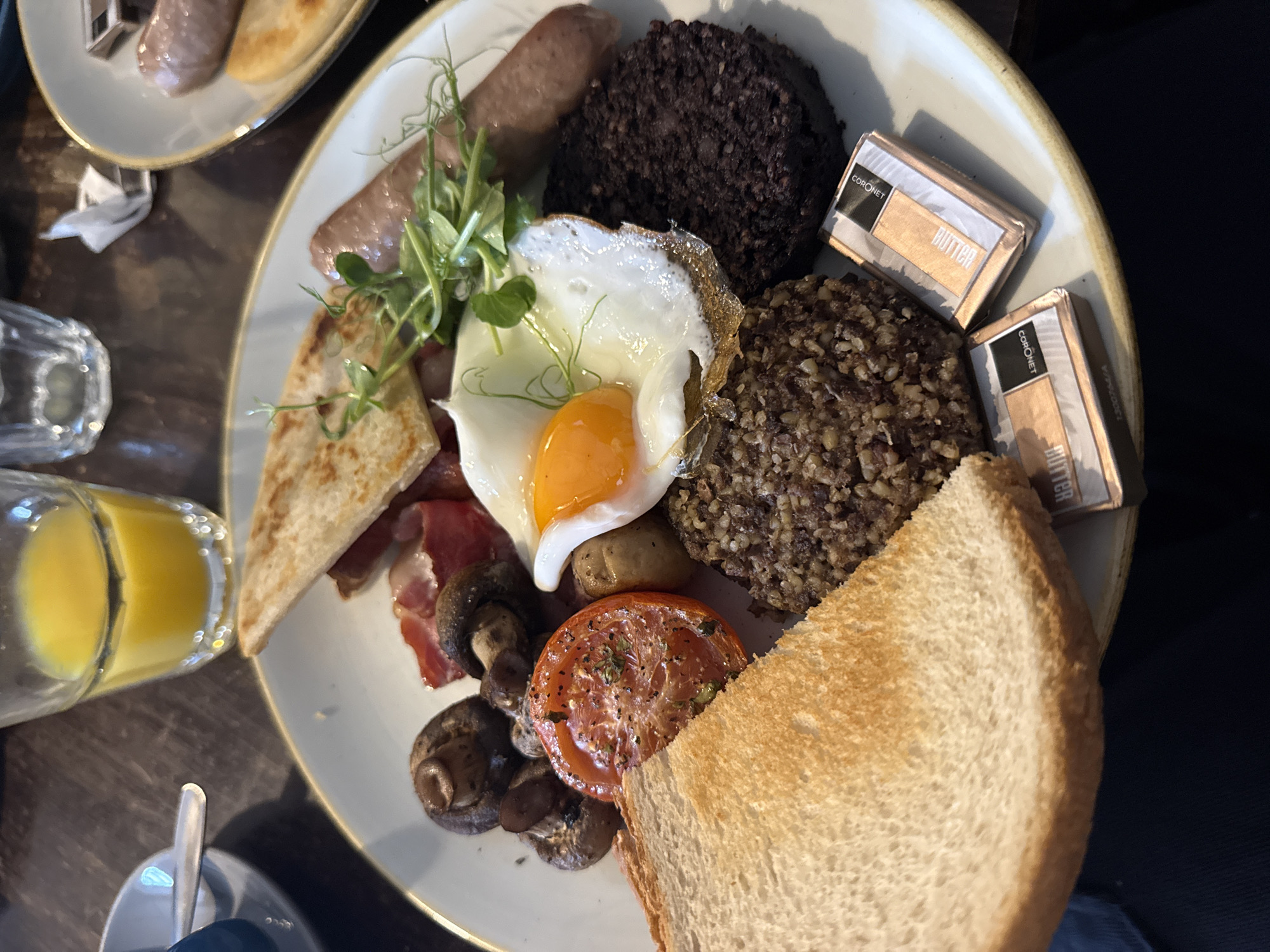The image shows a plate of food, a full Scottish breakfast, on a wooden table in an indoor setting. The plate contains sausage, black pudding, haggis, an egg, bacon, mushrooms, and a slice of toast. Two small packets of "Coronet Butter" are positioned on the plate. To the left, a glass of orange juice and a fork and spoon sit near the plate. Another plate containing sausage and what appears to be potato scones is visible in the background. The location is Unknown, Unknown.