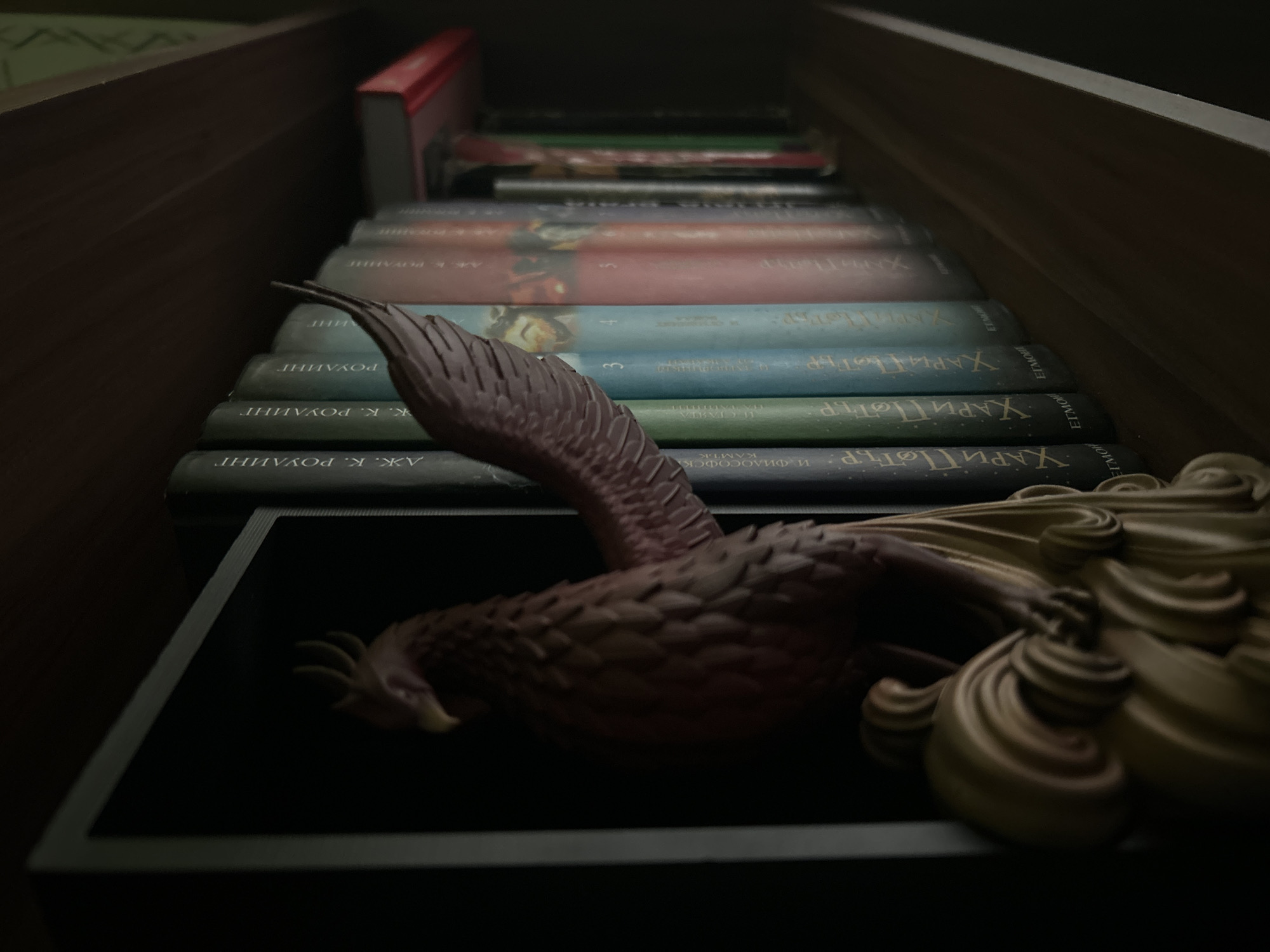 The image shows a collection of Harry Potter books, likely Russian translations, stacked on a dark shelf. A phoenix figurine, with dark reddish-brown body and lighter, ornate tail feathers, is prominently displayed in the foreground. The books appear to be in a dark, enclosed space, perhaps a bookshelf or a small cabinet. The focus is on the books and the figurine. There is no information to connect the location of the image to the city of Plovdiv, Bulgaria.