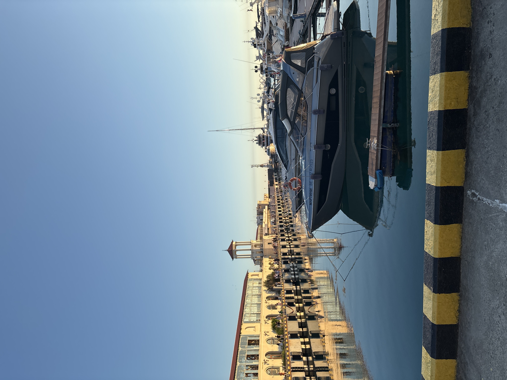 This is a photograph of a marina in Sochi, Russia, taken during the day. The scene is dominated by a clear blue sky and calm water reflecting the surrounding buildings and boats. Several yachts and boats are docked along a pier, with a prominent dark-colored motor yacht in the foreground. The pier has yellow and black striped bollards. In the background, a multi-story building with a red roof and numerous windows lines the waterfront, and a tower with a red roof stands out. The water is still, creating a mirrored image of the architecture and vessels. The overall atmosphere appears peaceful and serene. Text visible on the motor yacht reads "SUNSHINE 80" and "МАРТИН".