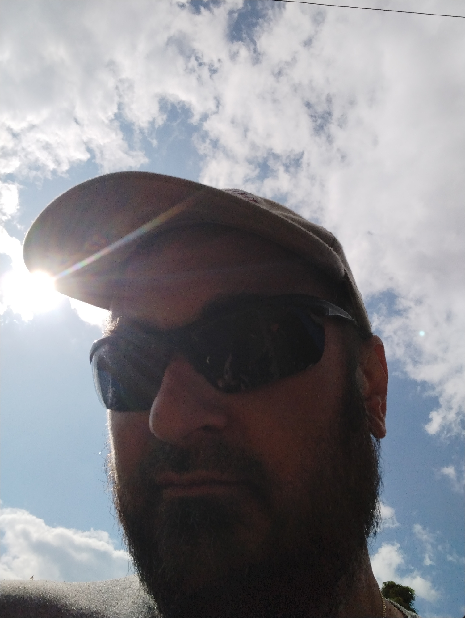 The image shows a close-up, low-angle shot of a person wearing a cap and sunglasses, looking upwards against a bright, cloudy sky. The sun is positioned to the left of the frame, creating a lens flare that partially obscures the cap and the person's face. The person has a beard and appears to be outdoors. The background consists of a blue sky with scattered white clouds. There is a thin black wire visible across the top of the frame. There are no discernible landmarks or elements that specifically indicate the location of Ширяево (ильинский), Russia.
