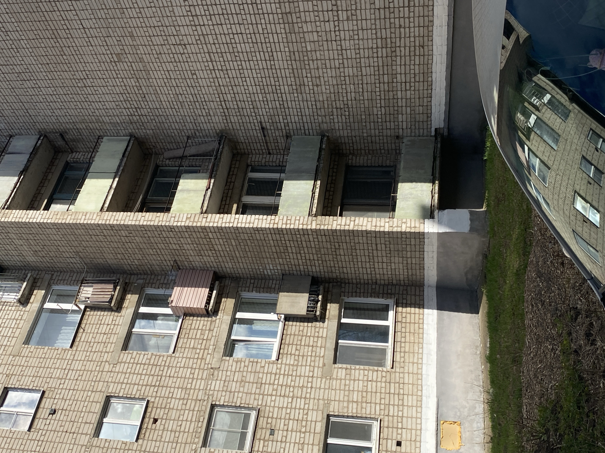 The image displays the exterior of a multi-story building, likely an apartment complex, under bright daylight conditions. The building features a light brown or tan brick facade with rows of windows and external fixtures.
On the upper floors, several recessed window sections are visible, some fitted with angled, light-colored covers, possibly for ventilation or shading. Lower down, the building features numerous rectangular windows with white frames; some appear open or partially open, reflecting the sky and clouds. Several windows on the mid-to-lower sections are accompanied by external air conditioning units, some covered by protective grates or housings, one with a distinctive beige/pinkish corrugated cover.
To the right of the building, a dark-colored car, possibly blue or black, is parked. Its highly reflective surface acts as a mirror, showing a distorted reflection of the building's facade, including windows and brickwork, and parts of the sky, suggesting another identical building might be reflected from the opposite side.
Adjacent to the building and alongside the car, there is a strip of green grass and some dry, sparse bushes lining a paved or concrete path. The base of the building shows areas where the brickwork is covered with white paint and exposed concrete or render. A small, square yellow object lies on the ground near the building's foundation.
The bright sunlight casts distinct shadows, indicating a clear, sunny day, likely late morning or early afternoon. No people or visible text are present in the static scene.