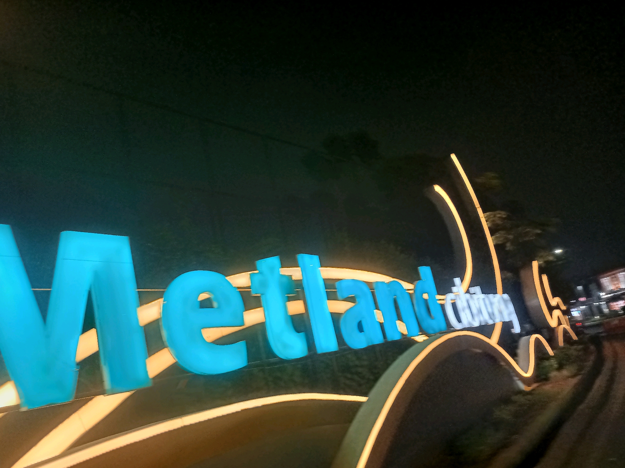 An illuminated blue sign spells out "Metland" in large capital letters against a dark background. The sign is outlined with a warm yellow light that follows the curve of a structure beneath it. To the right of the "Metland" sign, another word in white illuminated letters is partially visible, reading "city" and "tujuh". The overall scene appears to be a nighttime view of an entrance or landmark in Telagaasih, Indonesia, with a road curving to the right and blurred lights and structures in the background.