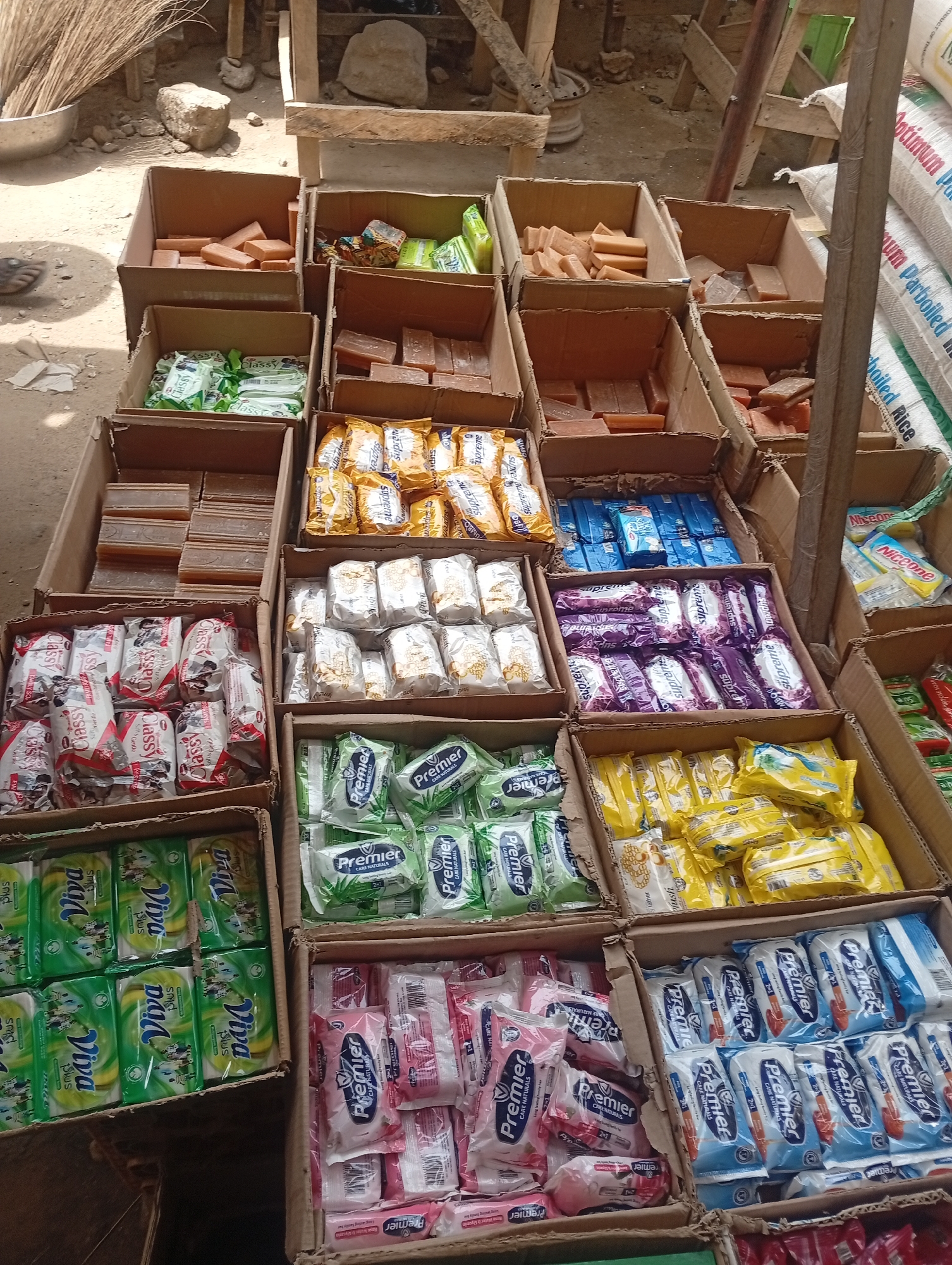The image shows a street-side vendor's display of various brands and types of soap in Sabon Tasha, Nigeria. The soaps are arranged in open cardboard boxes. The brands include "Viva Plus", "Classy", "Premier", and "Supreme", as well as plain bars of soap. Bags of rice are visible in the background. The setting appears to be an outdoor market.
