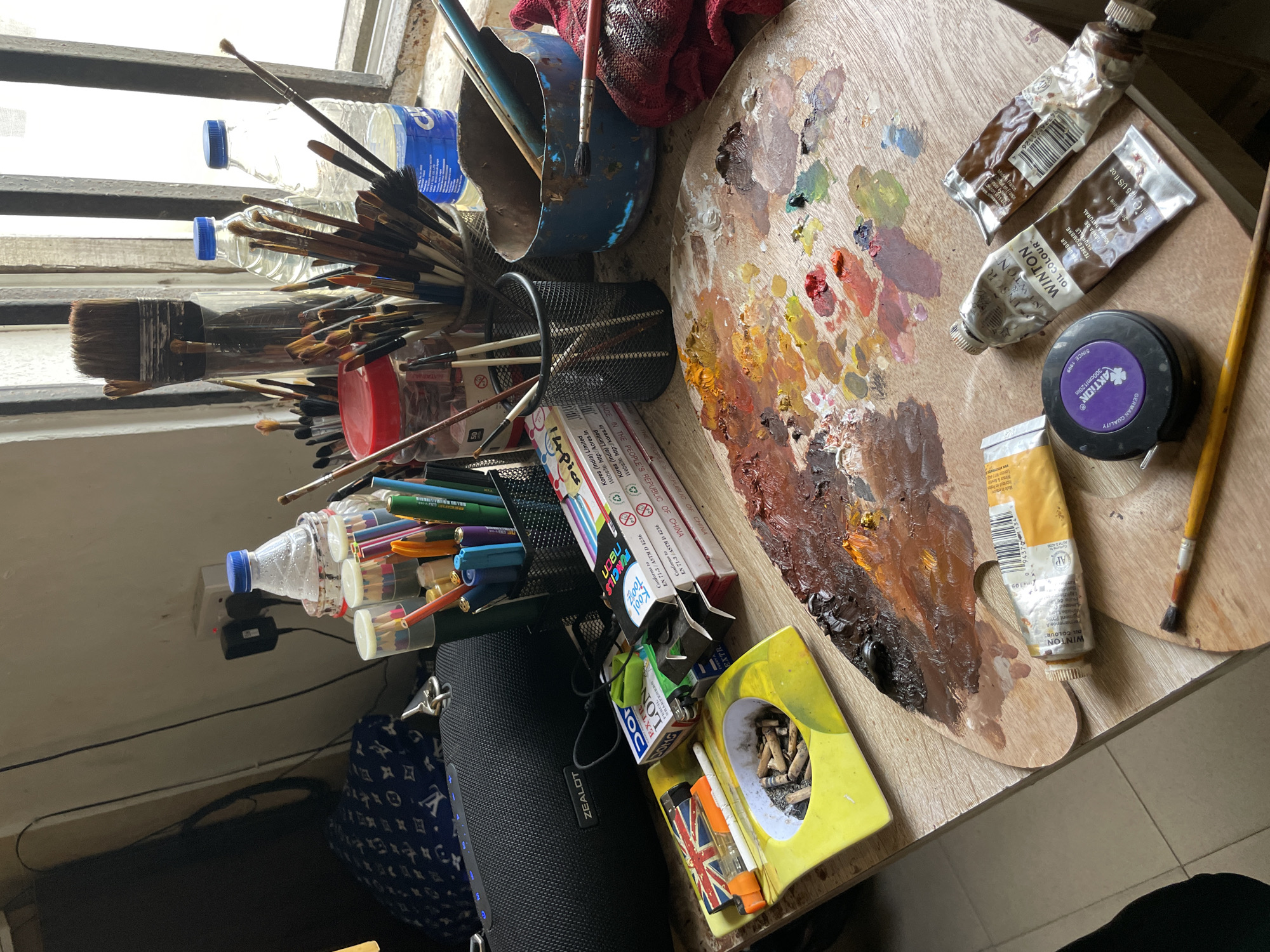 The image shows an artist's workspace. The scene includes an assortment of art supplies, such as paintbrushes in various containers, colored pencils, and a paint palette with a variety of oil paints. Tubes of Winton oil color are visible, in shades of brown and ochre, and one black round cylinder with GERMAN QUALITY. There's an ashtray containing cigarette butts and a lighter nearby. A Zealot speaker is placed beside the pencils. Two plastic water bottles can be seen in front of the window, together with a bucket full of brushes. The overall setting suggests an indoor environment. The city of Ado, Nigeria, is not explicitly present in the image's contents, but the location has been specified by the prompt.