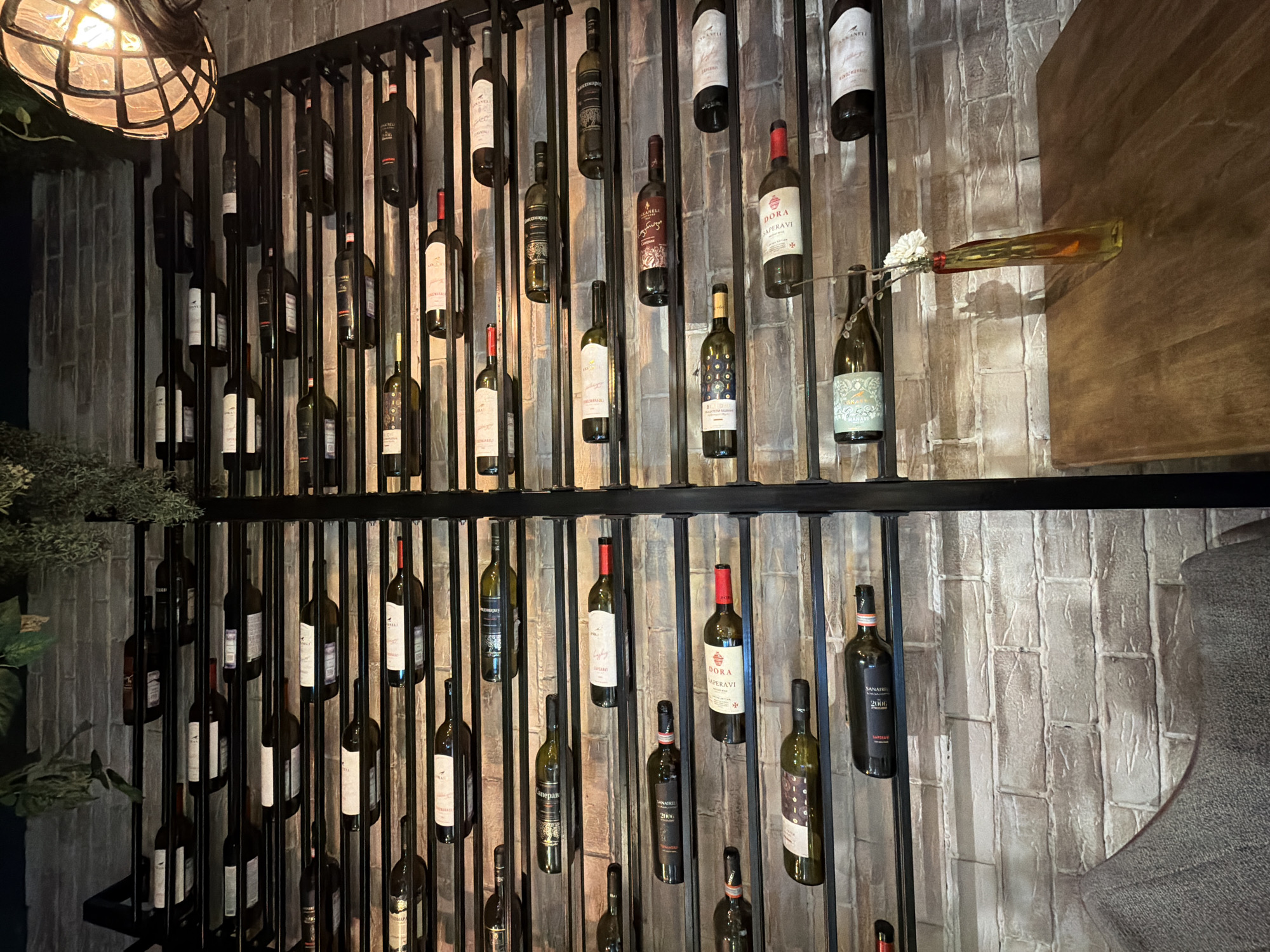 The image depicts a decorative wine rack against a textured brick wall. The rack is made of black metal and holds numerous wine bottles of various brands and colors, arranged in vertical rows. A light fixture hangs above the rack on the left. A decorative arrangement with white flowers and an amber bottle is present on the right side of the rack. A wooden surface is partially visible behind the rack on the right. The location is not specified in the image.