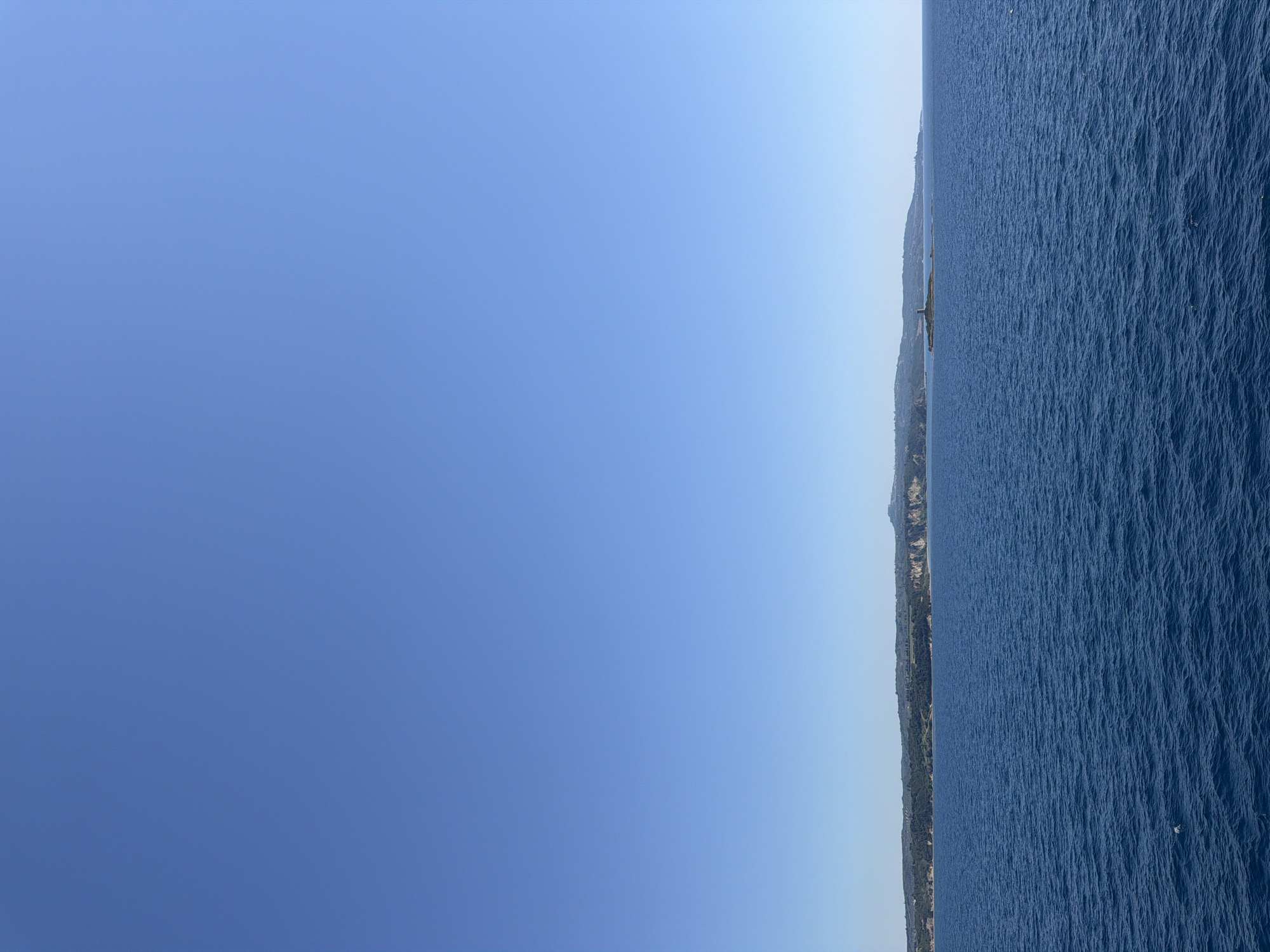 This is a photograph of the sea and the coast of the Municipal Unit of Kastro - Kyllini, Greece. The image captures a clear, blue sky above a vast expanse of deep blue water with gentle waves. On the horizon, a long, green coastline is visible, featuring rolling hills covered in vegetation and some cliffs. A small island with a prominent tower-like structure stands in the water off the coast. The overall scene depicts a serene coastal landscape under a bright, sunny sky.