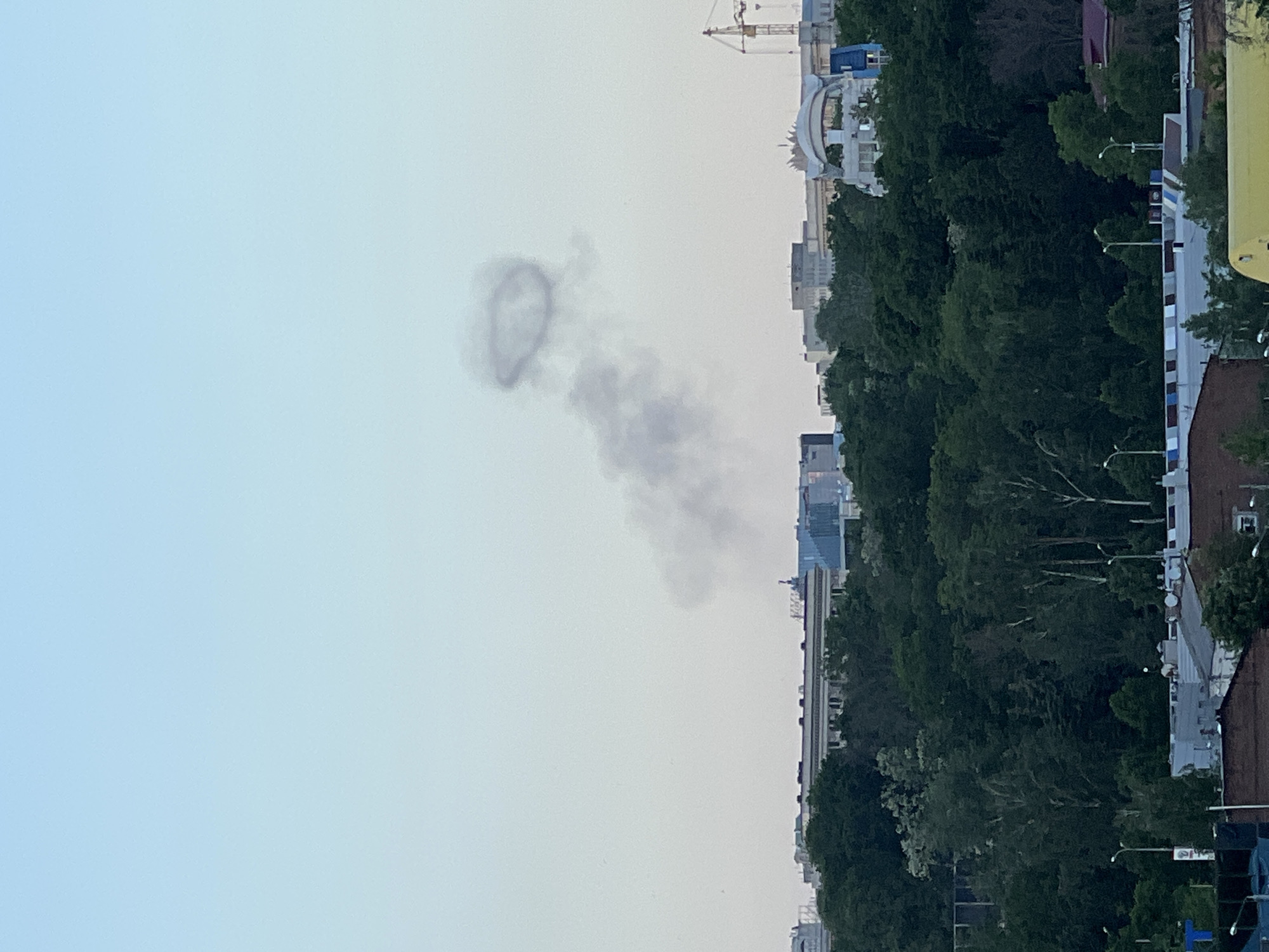 The image shows a column of gray smoke in the sky above a city. At the top of the smoke column, a circular shape is visible. The cityscape features a mix of buildings, including high-rises and low-rise structures, obscured partially by trees. A construction crane is visible in the background. The sky is a light blue-gray color.