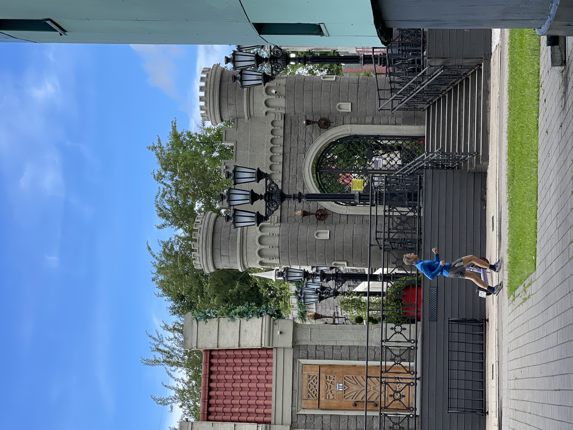The image depicts a man in mid-stride, wearing a blue long-sleeved shirt and grey shorts. He is on a paved walkway next to a grassy area, with a building resembling a castle in the background. The castle has stone walls, turrets, and ornate lamps. A red planter with greenery is visible near the castle entrance. The sky is blue with scattered clouds, suggesting it is daytime with fair weather. The architectural style of the building, particularly the castle-like features and the overall aesthetic, might suggest a theme park or a specially designed building in a public space. The man appears to be engaged in some form of exercise or recreational activity, possibly jogging or walking briskly. The scene is set outdoors in what appears to be a well-maintained urban or suburban area. No visible text is present in the image.