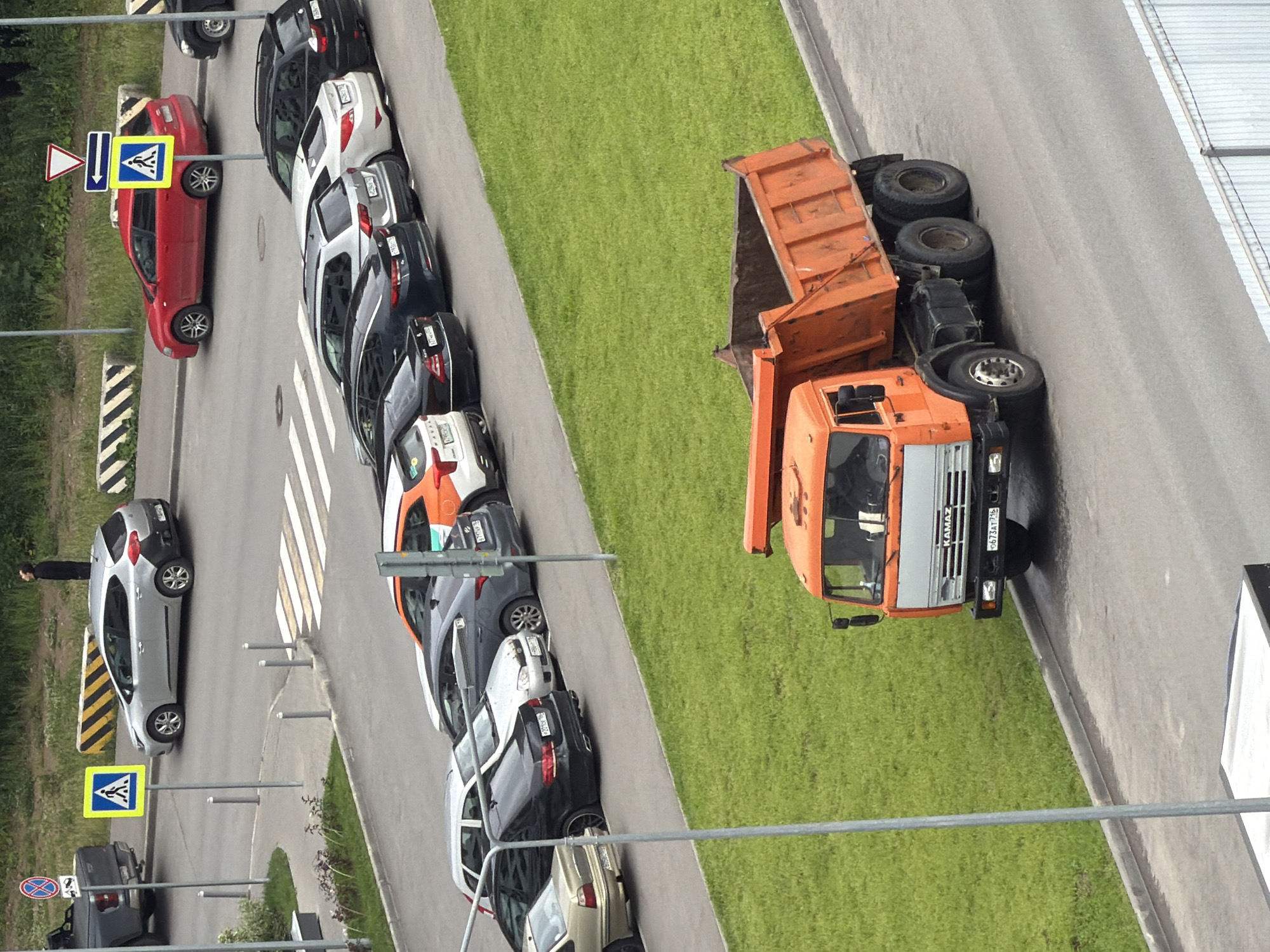 An outdoor, high-angle medium shot shows a street scene in Unknown, Unknown, with a focus on parked cars and a dump truck. A silver Kia sedan is parked on the street, facing right, with a pedestrian crossing sign and a roundabout sign next to it. A red Kia hatchback is parked to the right of the silver Kia. Several other cars are parked in a row further down the street, including a white Kia, a gray Kia, and a black Kia. In the foreground, an orange Kamaz dump truck is parked, facing left. The truck has a weathered appearance and its bed is raised. The street is wet, suggesting recent rain, and a patch of green grass is visible on the right side of the frame. A person is visible standing on a sidewalk in the background, looking up.