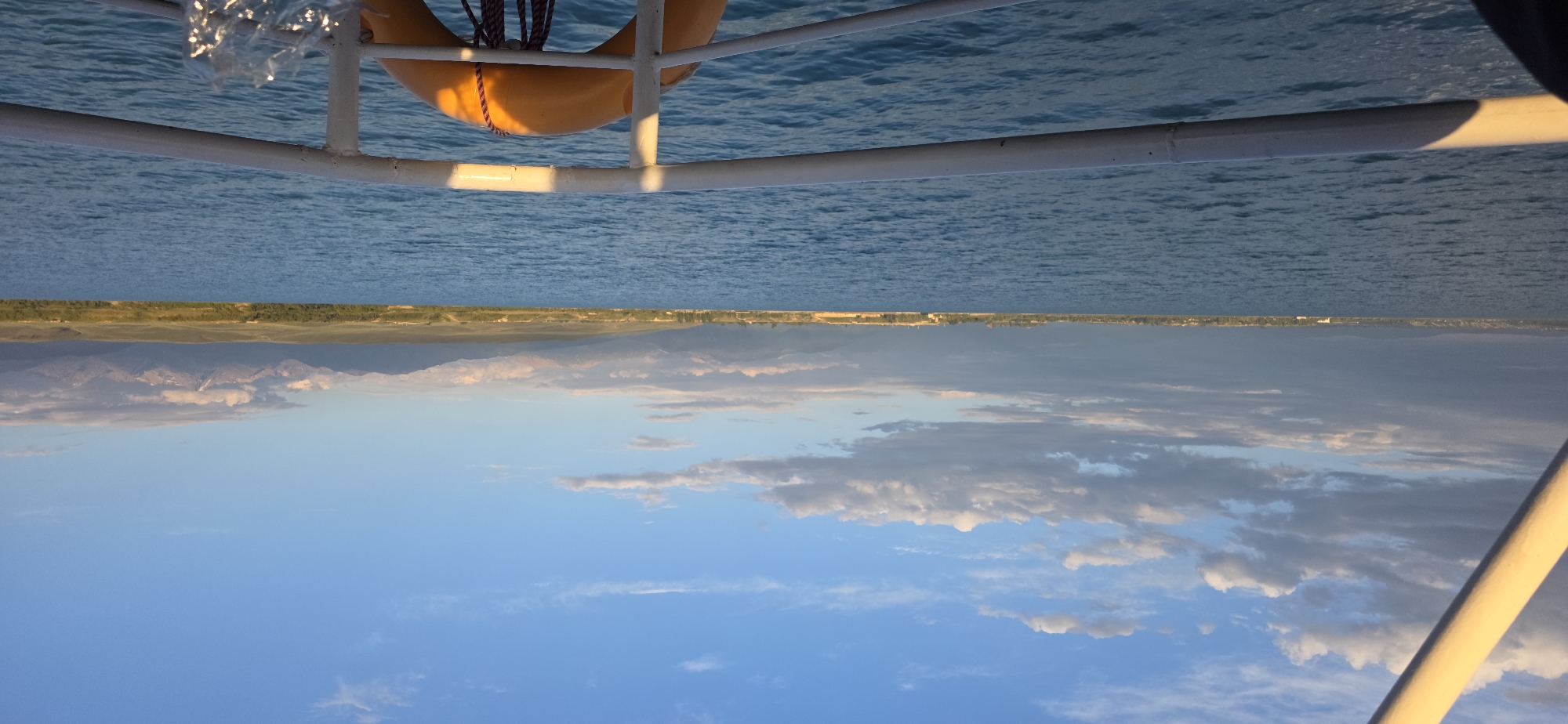 The image is an upside-down, wide-angle shot of a body of water with a distant shoreline and mountains. In the foreground, a white railing with a yellow life preserver attached is visible. The water is a vibrant blue with ripples, suggesting movement. Beyond the water, a green and brown shoreline stretches across the horizon, backed by a range of mountains that appear snow-capped in places. The sky above is a clear blue, dotted with scattered white clouds, particularly towards the horizon where they blend with the mountain range. The lighting suggests it is either early morning or late afternoon, as the sun casts a warm glow on the railing and highlights the clouds. The overall scene evokes a sense of tranquility and natural beauty. The image is a photograph taken from a boat, as indicated by the railing and the perspective. There is no visible text in the image.