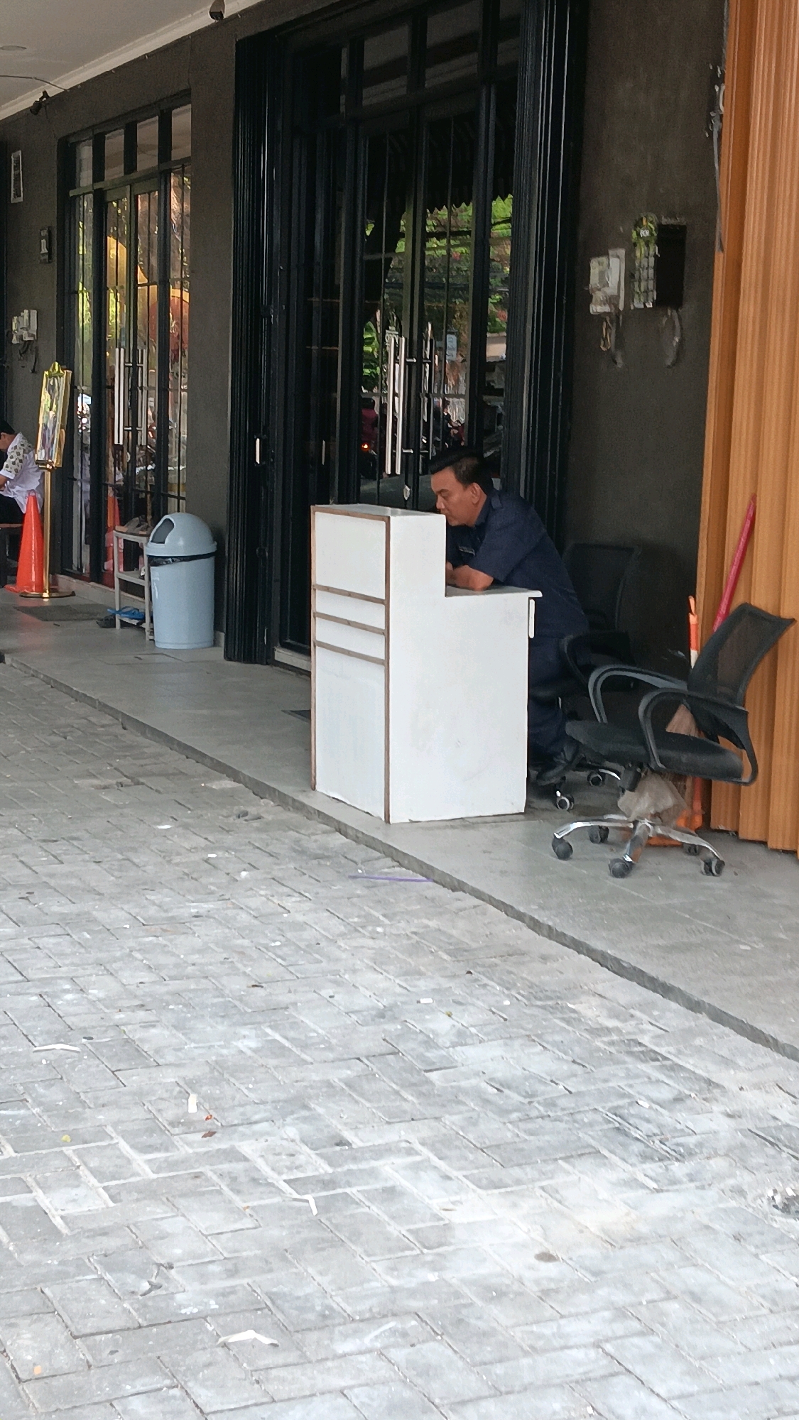 A man in a dark blue uniform, possibly a security guard, leans on a white reception desk with wooden trim. He is positioned to the right of the desk, facing inwards. Behind him, two black office chairs with wheels are visible. The scene takes place outdoors, on a paved walkway with a herringbone brick pattern. To the left of the desk, an orange traffic cone and a grey trash can are present. The background features a building with large, dark-framed glass doors and windows. The overall environment suggests a commercial or public space in an urban setting, possibly in South Jakarta, Indonesia, given the context.