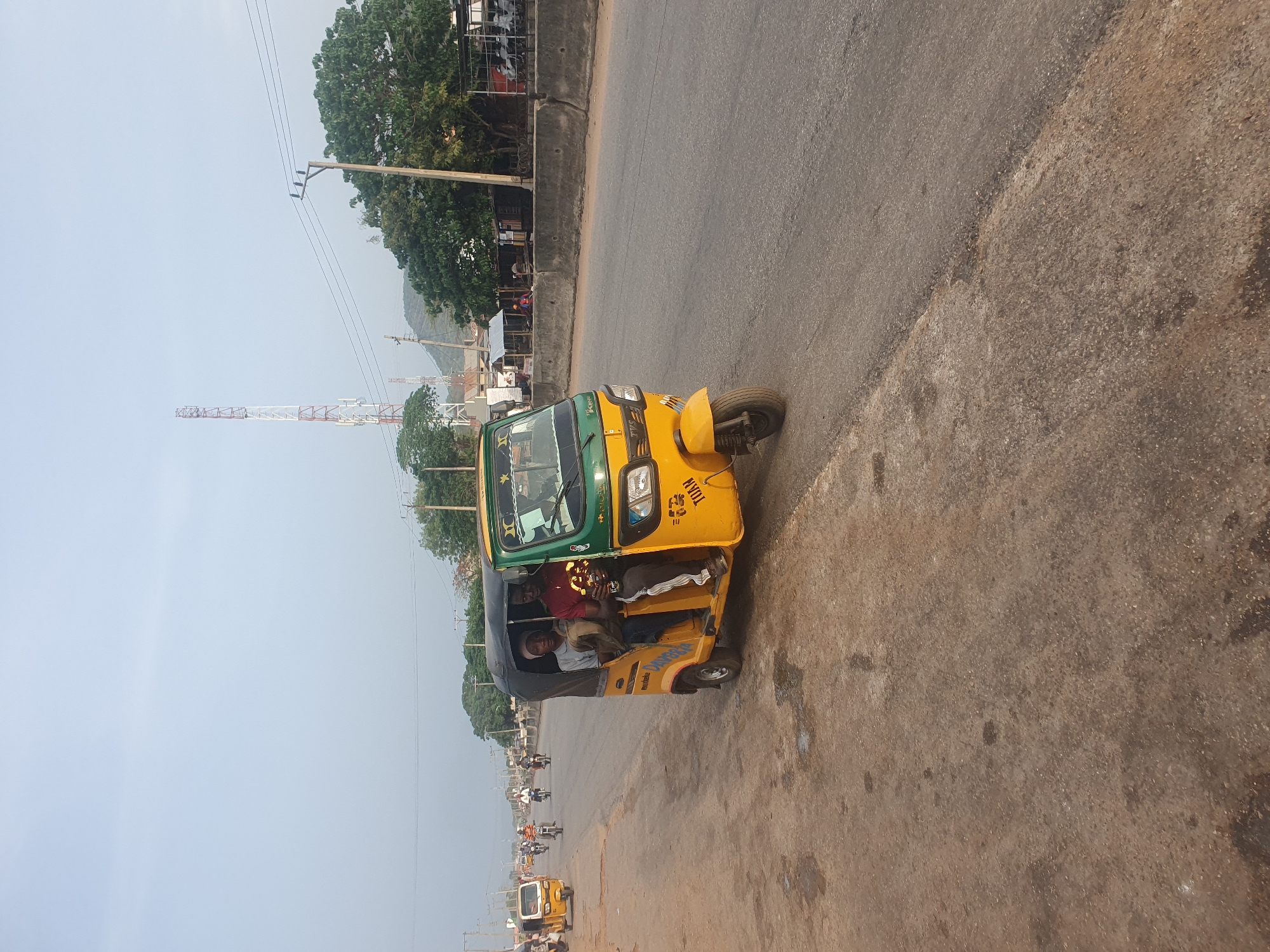 The image depicts a yellow and green commercial tricycle, commonly known as a "keke," in Lokoja, Nigeria. Two passengers are visible inside the tricycle. The tricycle is parked on the side of a paved road. In the background, a red and white communication tower is visible. Power lines run along the road. Additional tricycles and motorcycles are on the road in the distance. Trees and a building line the road in the background.