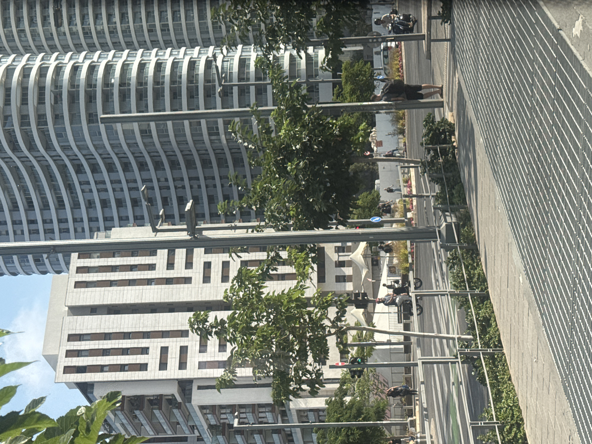 The image captures a street scene in Tel Aviv, Israel, with a focus on urban architecture and pedestrian activity. Prominently featured are modern, tall buildings with distinctive curved facades on the right and a more rectilinear, lighter-colored building with darker vertical accents on the left. The foreground shows a wide, paved walkway with a metal grate running diagonally across it, bordered by a metal railing and a dense row of green bushes. Beyond the walkway, a street is visible with a pedestrian crossing and traffic lights.
Several people are present in the scene. On the left side of the street, a person is walking, and further down, a person is riding a bicycle. Near the center, a person is seated on a bench, seemingly on an electric scooter or small vehicle, while another person stands nearby. On the right side of the street, a person is standing on the sidewalk with a backpack, and a person is riding a motorcycle. The environment is sunny with clear blue skies, indicating a daytime setting. Trees with green foliage are interspersed throughout the scene, adding a touch of nature to the urban landscape. The overall impression is a bustling city street with a mix of residential and commercial structures.