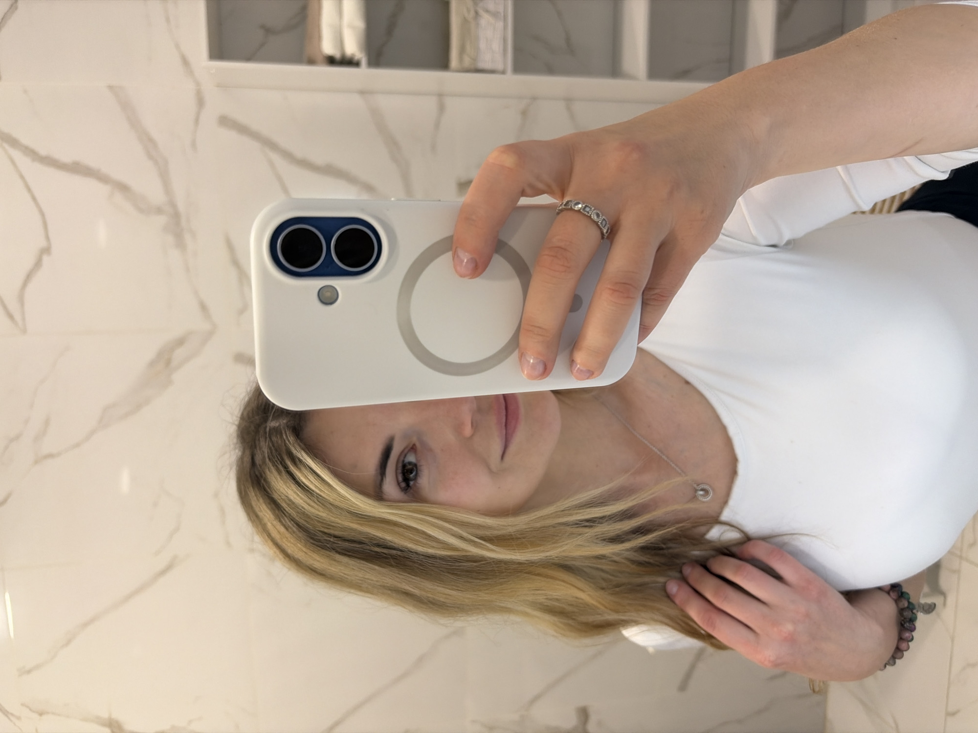 A woman is taking a selfie in a bathroom with white marble walls. The bathroom has some decorative items in the background. The woman is holding a white smartphone in her right hand, and she's wearing a ring on her ring finger. She has blonde hair, fair skin, and is wearing a white top. She is also wearing a necklace. The woman is indoors. It is not possible to identify the exact location in the city of Budva, Montenegro.