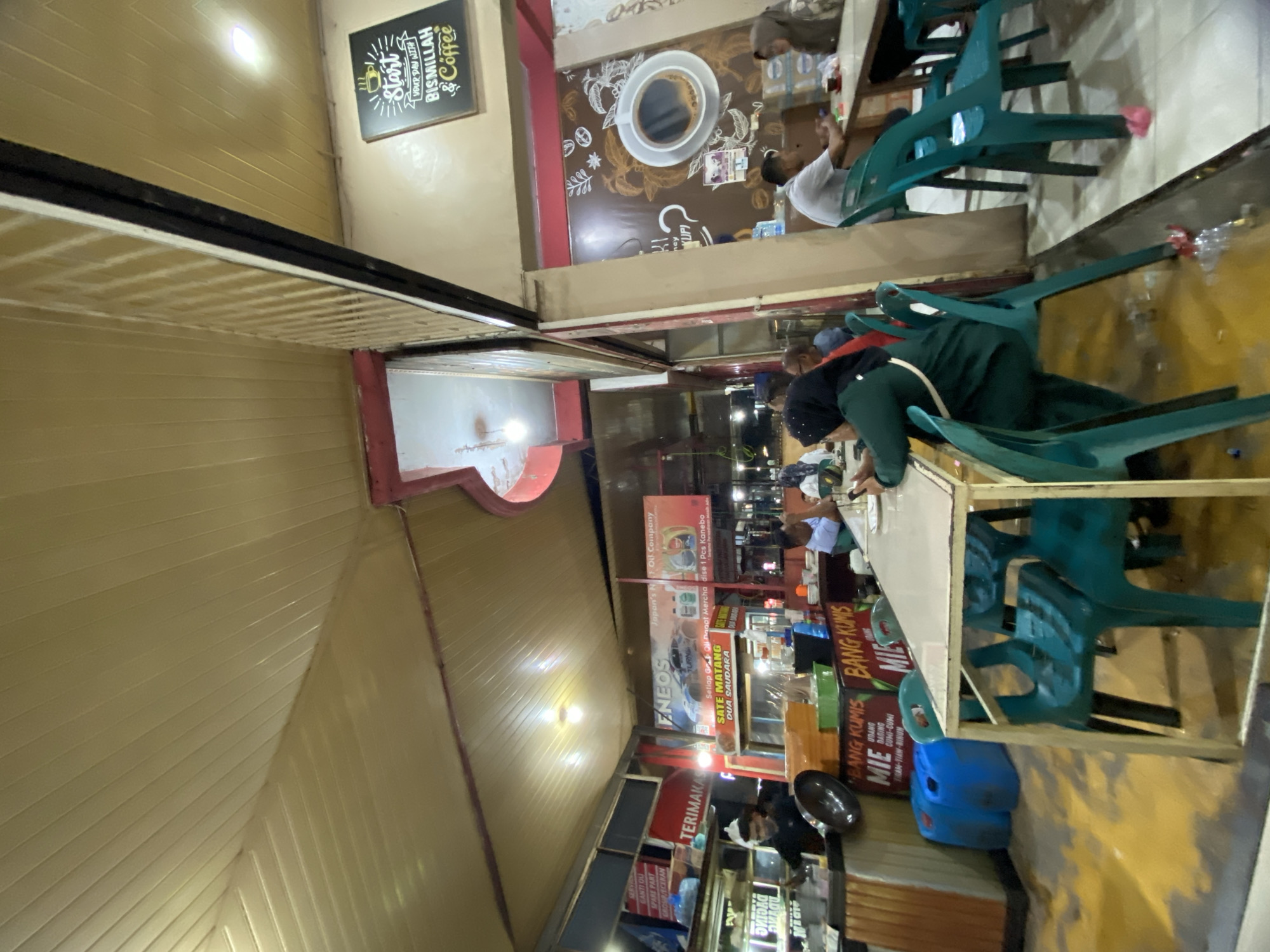 The image depicts an outdoor dining area at a food stall in Lhokseumawe, Indonesia. The stall appears to be a popular spot, with several patrons seated at tables and a counter visible where food is being prepared. The atmosphere is casual and communal.
In the foreground, a person in a green hijab sits at a table, looking towards the right side of the frame. They are wearing a dark green outfit. Next to them, an empty green plastic chair is positioned at the table, which has a light-colored top and yellow metal legs. Another green plastic chair is visible behind the first person, and more chairs are scattered around the seating area. The floor is tiled and appears somewhat wet, with some debris, possibly from spilled drinks or food, scattered around.
To the left, a food stall counter is visible, with various containers and equipment for food preparation. A person in a dark shirt is working behind the counter, appearing to serve or prepare food. Blue plastic barrels are placed on the ground near the counter. A banner with text, partially obscured, is mounted above the counter, and another banner featuring an advertisement for Eneos oil and "Sate Matang Dua Saudara" is prominently displayed.
Further back, more tables and chairs are set up, occupied by patrons. The scene is illuminated by overhead lights, suggesting it is evening or night. A sign on the wall to the right features a graphic of a coffee cup with text including "Start Your Day With Bismillah & Coffee." The ceiling is made of pale yellow corrugated panels. The overall environment suggests a lively, bustling eatery catering to local residents.