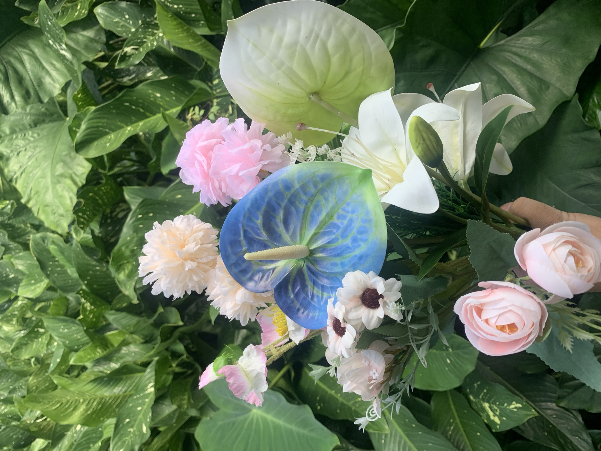An artificial flower arrangement is held in a hand, positioned in front of a lush, green leafy background. The arrangement includes a prominent blue and green anthurium flower, a white lily, several pink roses, and smaller white and pink flowers, along with decorative foliage. The background appears to be an outdoor setting with large, glossy green leaves, possibly in a garden or park. There is no indication of the specific location in Kuala Pembuang, Indonesia. The image is focused on the floral display, with the hand holding the bouquet visible at the bottom right.