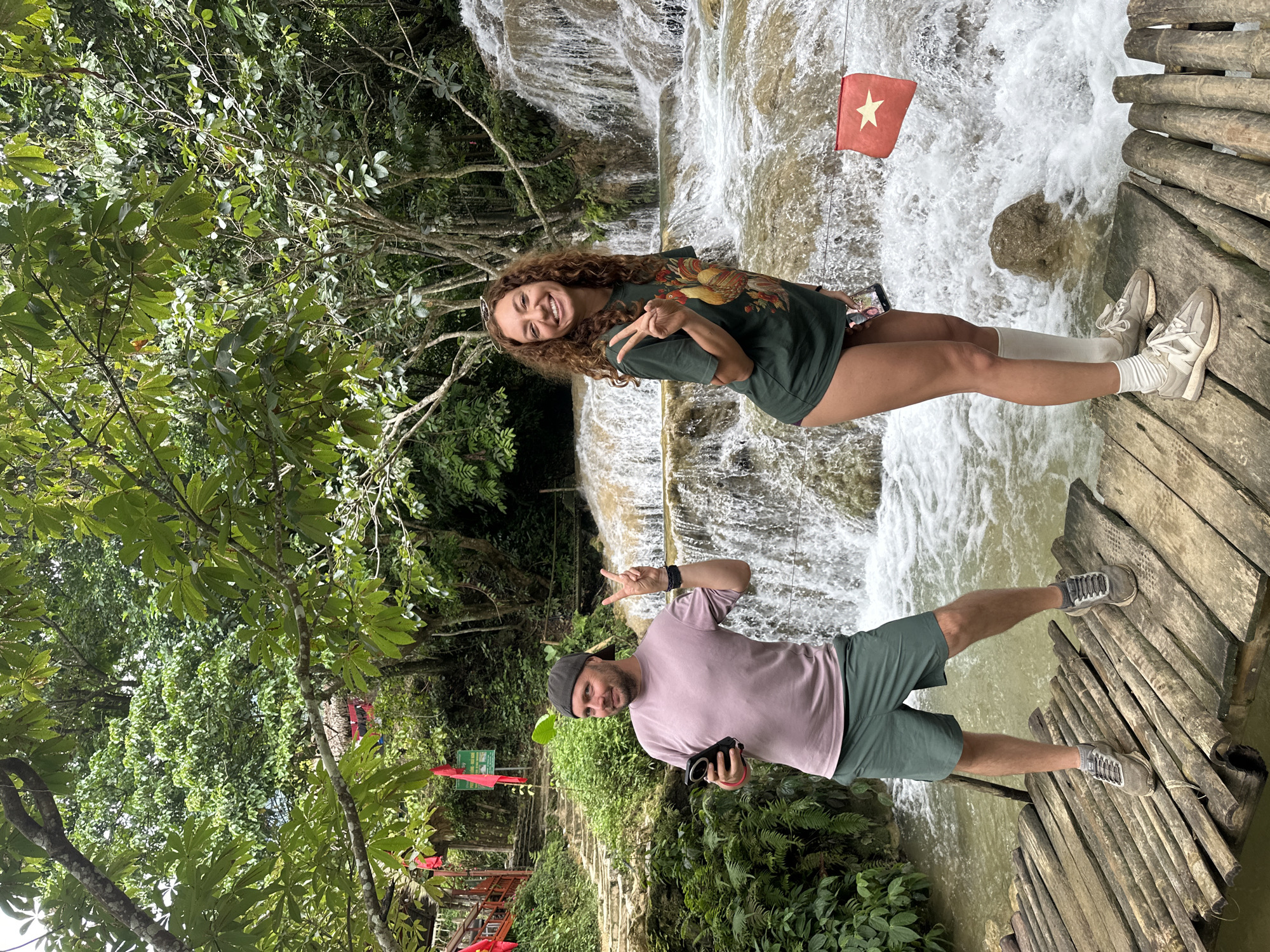 Two individuals, a man and a woman, are standing on a wooden boardwalk in front of a cascading waterfall. The man, on the left, is wearing a purple t-shirt and green shorts, and has a dark cap on. He holds a black smartphone in his left hand and is making a peace sign with his right hand. The woman, on the right, is wearing a green t-shirt and appears to be wearing shorts or a skirt. She is also making a peace sign with her right hand and holds a smartphone in her left hand. A red flag with a yellow star, likely the Vietnamese flag, is visible to the right of the woman, fluttering in the breeze. The environment is lush and green, with dense vegetation and trees surrounding the waterfall and boardwalk. The waterfall appears to be a natural rock formation with multiple tiers. The presence of a Vietnamese flag suggests the location is in Vietnam, specifically Cổ Lũng Commune.