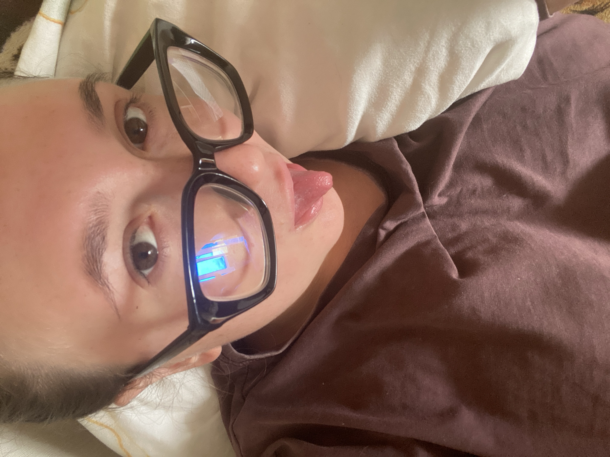 The media file shows a close-up, rotated shot of a person wearing black-rimmed glasses. The person is lying down, possibly on a bed, with a white pillow visible behind their head. They are wearing a dark-colored shirt. The person's tongue is slightly extended, and their mouth is open. The glasses have blue light reflections on the lenses, suggesting the presence of a screen or light source. The overall mood seems casual and perhaps playful. The image is not geographically specific, beyond the provided context of Chaikovsky, Russia, and there is no visible text.
