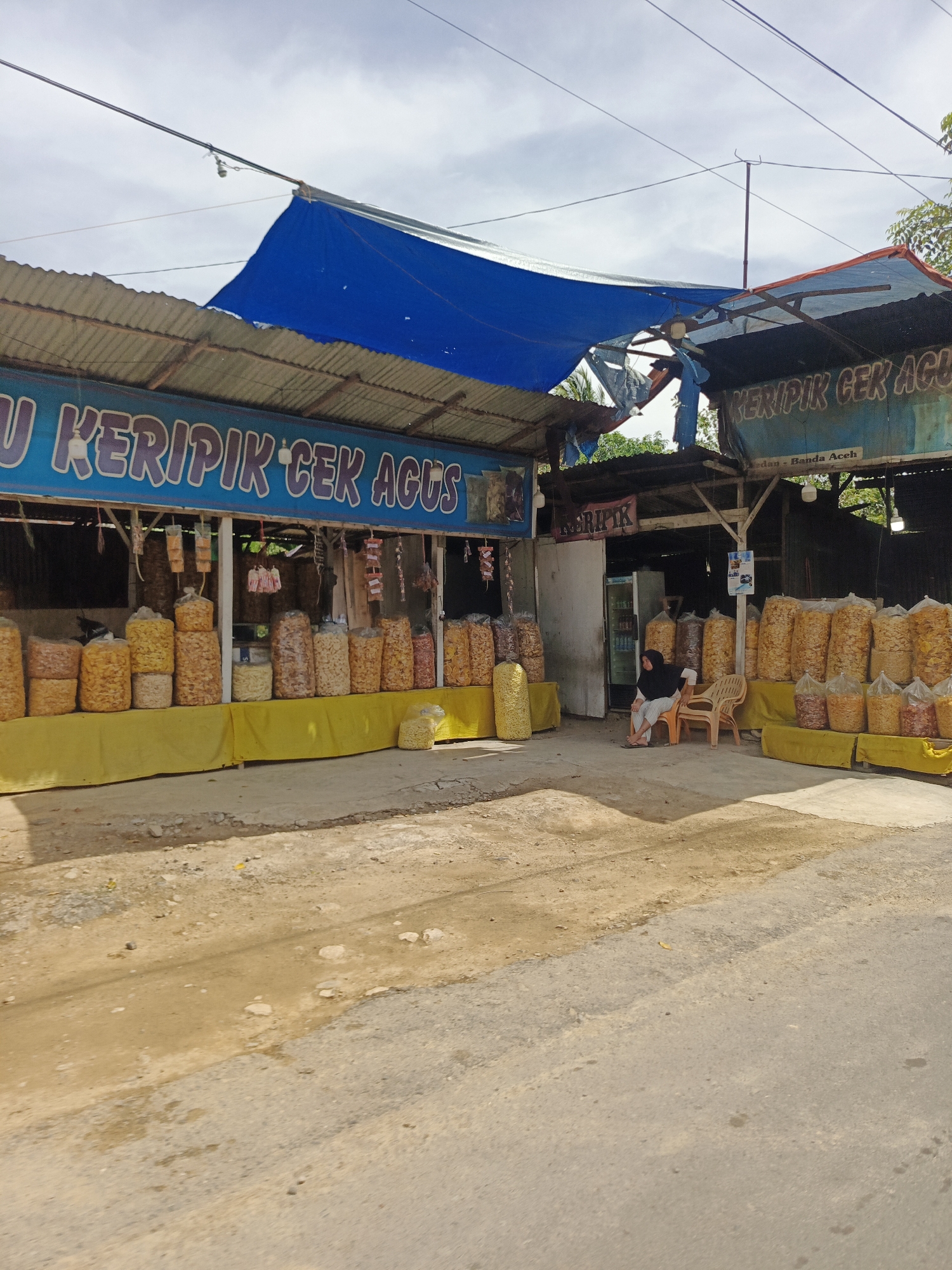 The image depicts an outdoor food stall in Sawang, Indonesia, specializing in "keripik" (chips). The stall features a long display counter covered with yellow fabric, showcasing numerous large plastic bags filled with various types of chips. Behind the counter, a blue tarp and corrugated metal sheeting provide shelter for the stall. A sign with blue lettering reads "U KERIPIK CEK AGUS," indicating the name of the establishment.
In the middle section of the stall, a woman wearing a black headscarf and a dark-colored dress is seated on a plastic chair, facing towards the right of the image. Two other plastic chairs are visible nearby. A refrigerator is also present behind the counter. The ground in front of the stall is a mix of dirt and asphalt, with some debris scattered around. The overall scene suggests a typical roadside vendor in a tropical environment.