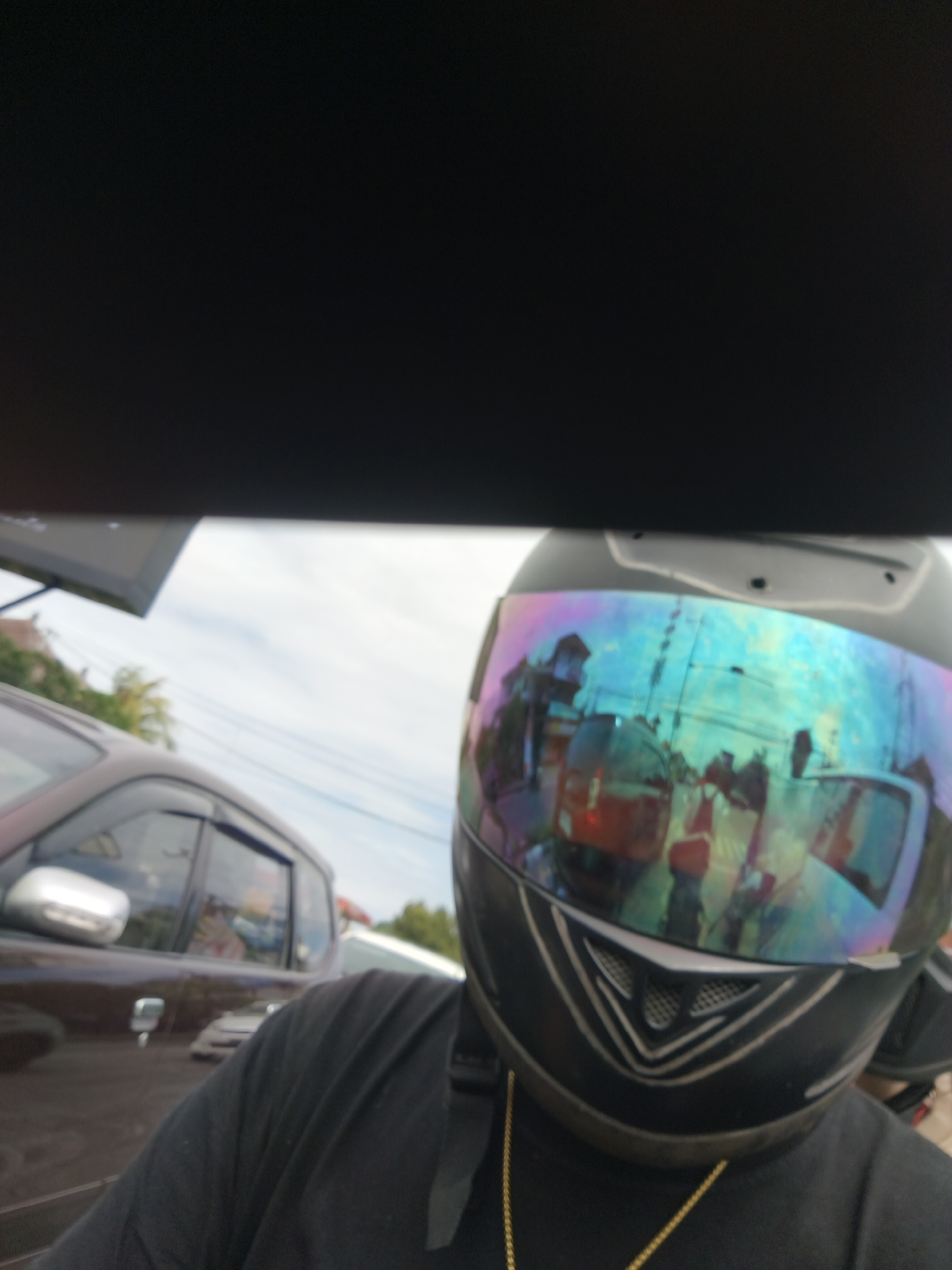 In Denpasar, Indonesia, a person wearing a black motorcycle helmet with a reflective, iridescent visor is shown. The visor displays distorted reflections of a street scene, including buildings, other vehicles, and people. In the foreground, to the left of the helmet, a portion of a dark-colored car is visible. The background includes greenery and a cloudy sky. The image appears to be taken from the perspective of the motorcycle rider.