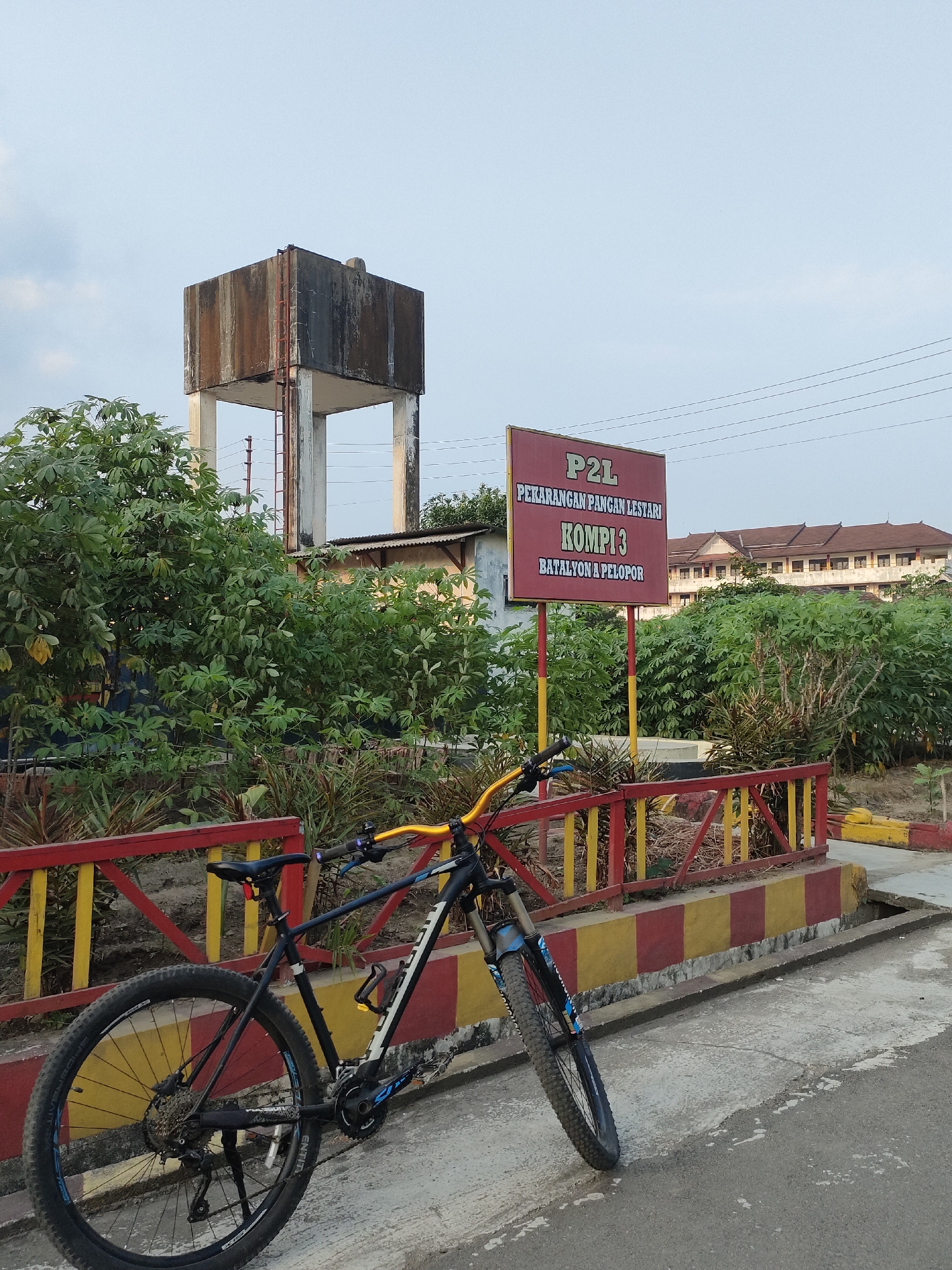 The image captures a scene in Banguntapan, Indonesia, featuring a black mountain bike parked beside a red and yellow striped barrier. Behind the barrier, lush green vegetation surrounds a tall, weathered concrete water tower. To the right of the water tower, a red sign with white and yellow lettering reads "P2L PEKARANGAN PANGAN LESTARI KOMPI 3 BATALYON A PELOPOR." Further in the background, a multi-story building with a brown tiled roof is visible, set against a pale sky. The ground is a mix of concrete and asphalt, with some patches of wear and tear.