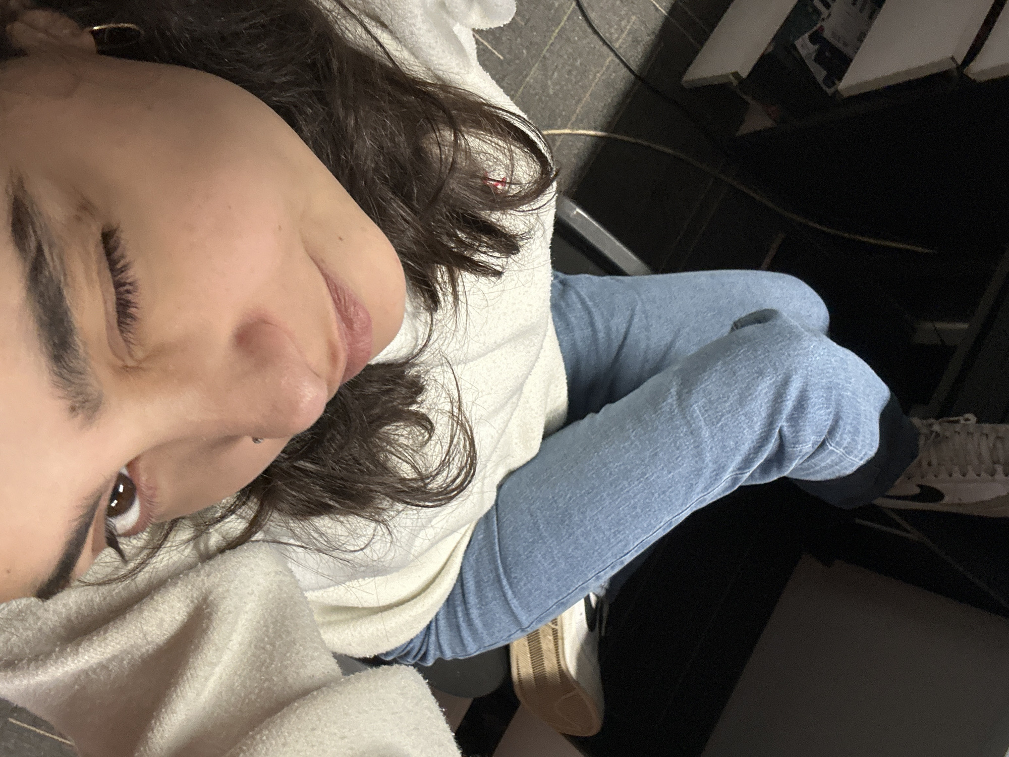 A woman sits in an interior setting. She has brown hair, brown eyes, and is wearing a hoop earring and jeans. She's wearing a white sweater and what appears to be a white towel. The location can't be determined from the given image.