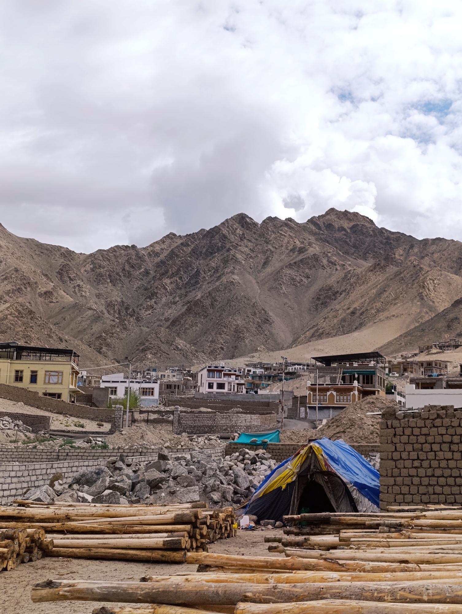 In Leh, India, a mountain village scene unfolds under a cloudy sky. The foreground is dominated by stacks of freshly cut logs, some neatly piled and others scattered, suggesting ongoing construction or renovation. Among these logs, a makeshift tent with blue, yellow, and black tarps provides shelter. Behind the logs and tent, a variety of buildings are visible, ranging from simple brick structures to more elaborate multi-story houses, indicative of a developing urban area. The backdrop is a dramatic mountain range, its rugged slopes covered in scree and sparse vegetation, characteristic of the arid Himalayan landscape. The overall impression is one of a remote, mountainous settlement actively engaged in development, with a blend of natural grandeur and human activity.
