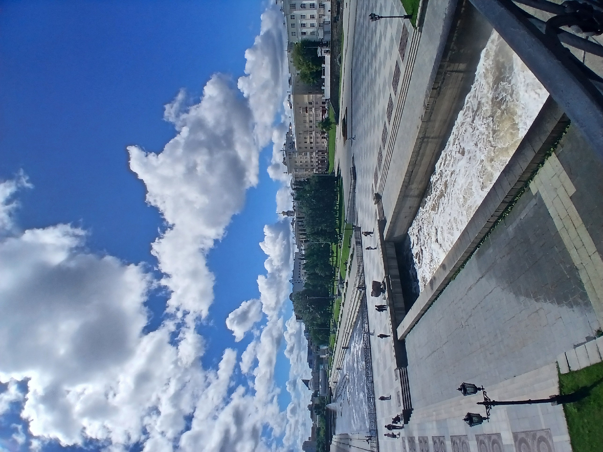 The image depicts a sunny day with scattered clouds over the Iset River embankment in Yekaterinburg, Russia. The embankment is paved with large square tiles and lined with trees and buildings. Several people are visible strolling along the embankment. A wide, turbulent river flows through a concrete channel, with a fountain visible in the distance. The scene suggests a public gathering space in an urban environment.